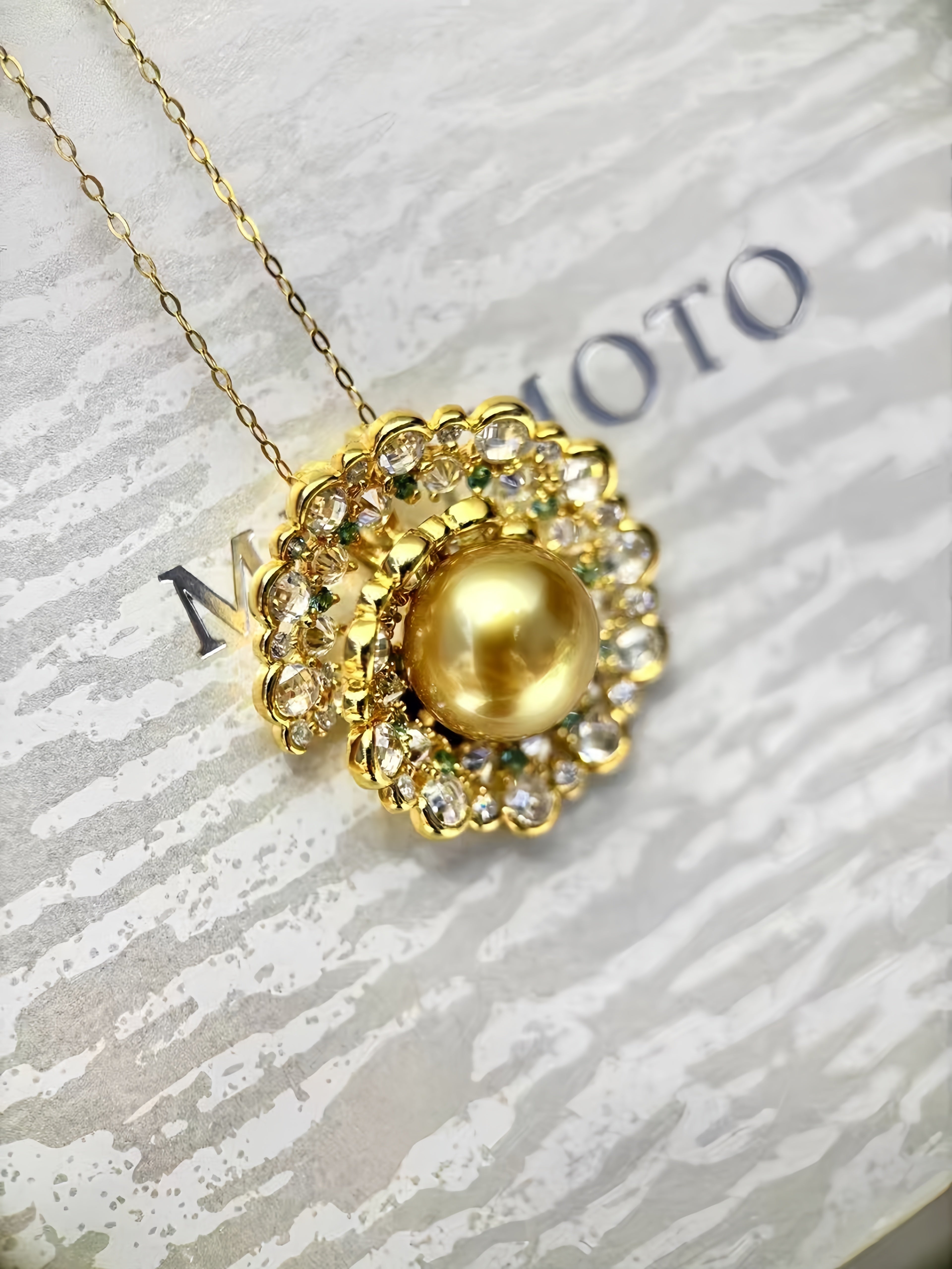 A gold pendant necklace featuring a large round gold pearl surrounded by a halo of clear rhinestones, resting on a textured white surface with a gold chain extending off the top left corner.