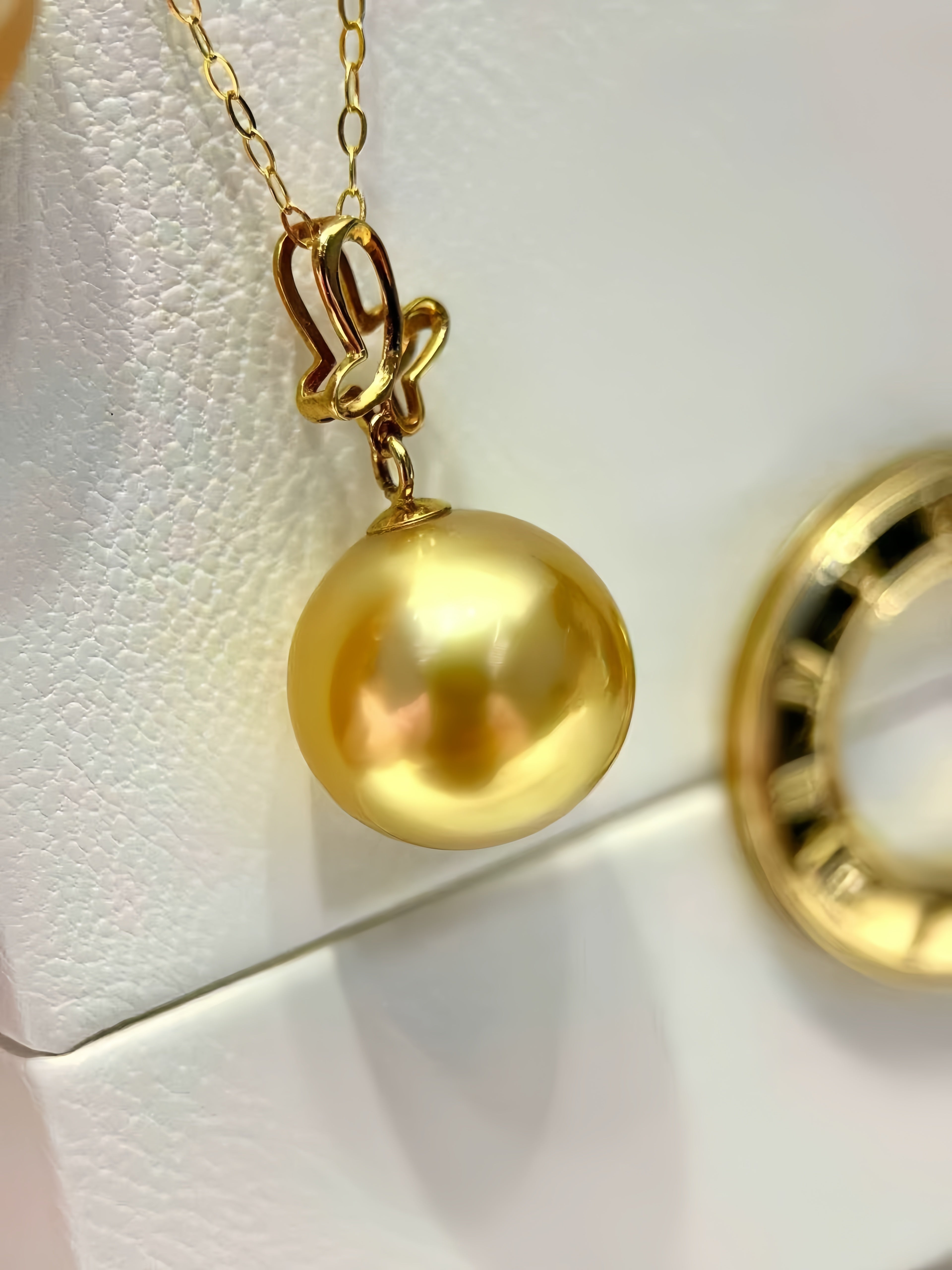 A gold pendant necklace featuring a large, polished round pearl-like drop and an ornate curved bail, displayed on a textured white surface.