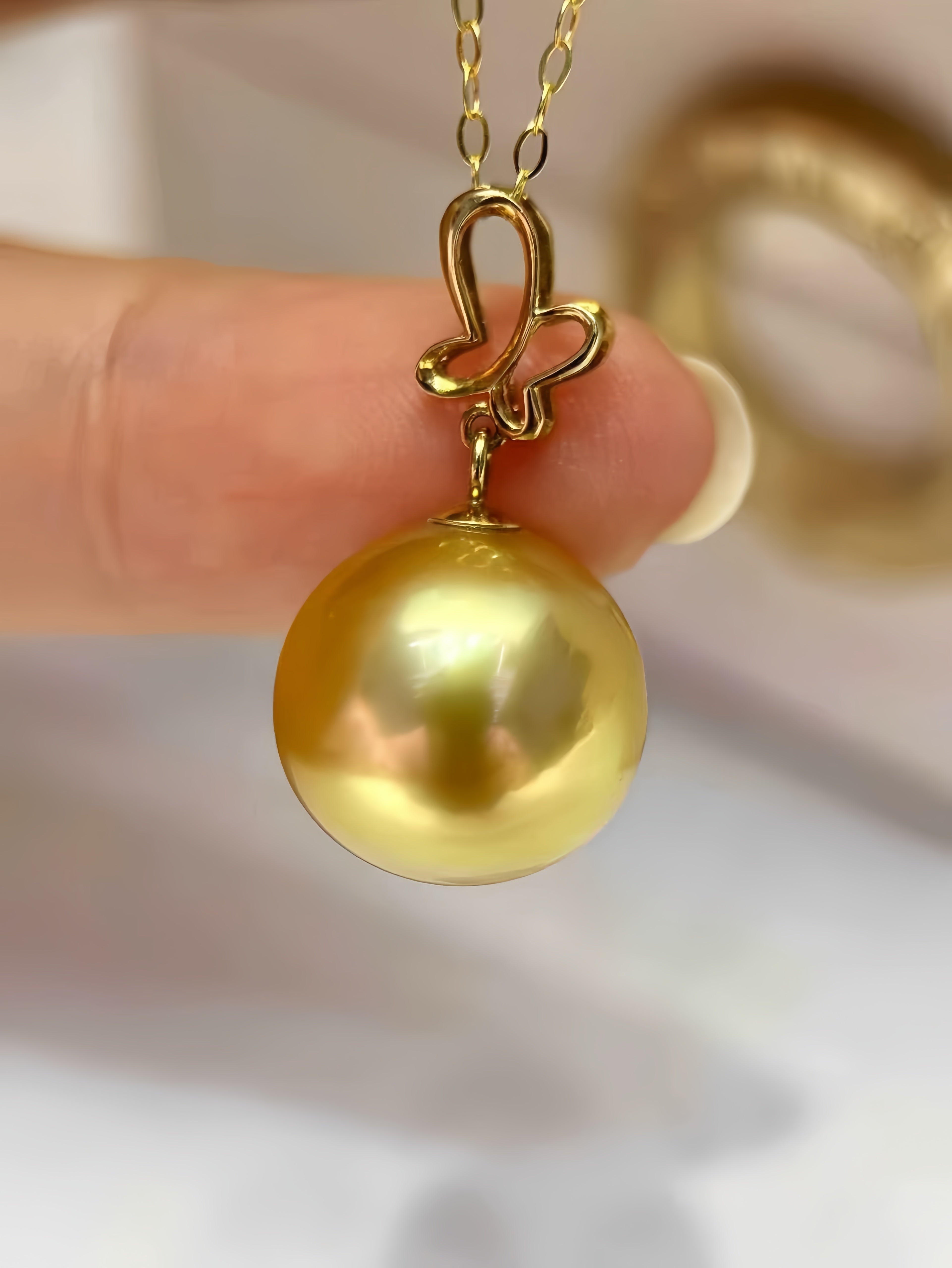A close-up of a gold pendant necklace featuring a large, round, lustrous yellow gold pearl suspended from a decorative gold bail, held by a person's fingers against a soft, out-of-focus light background.