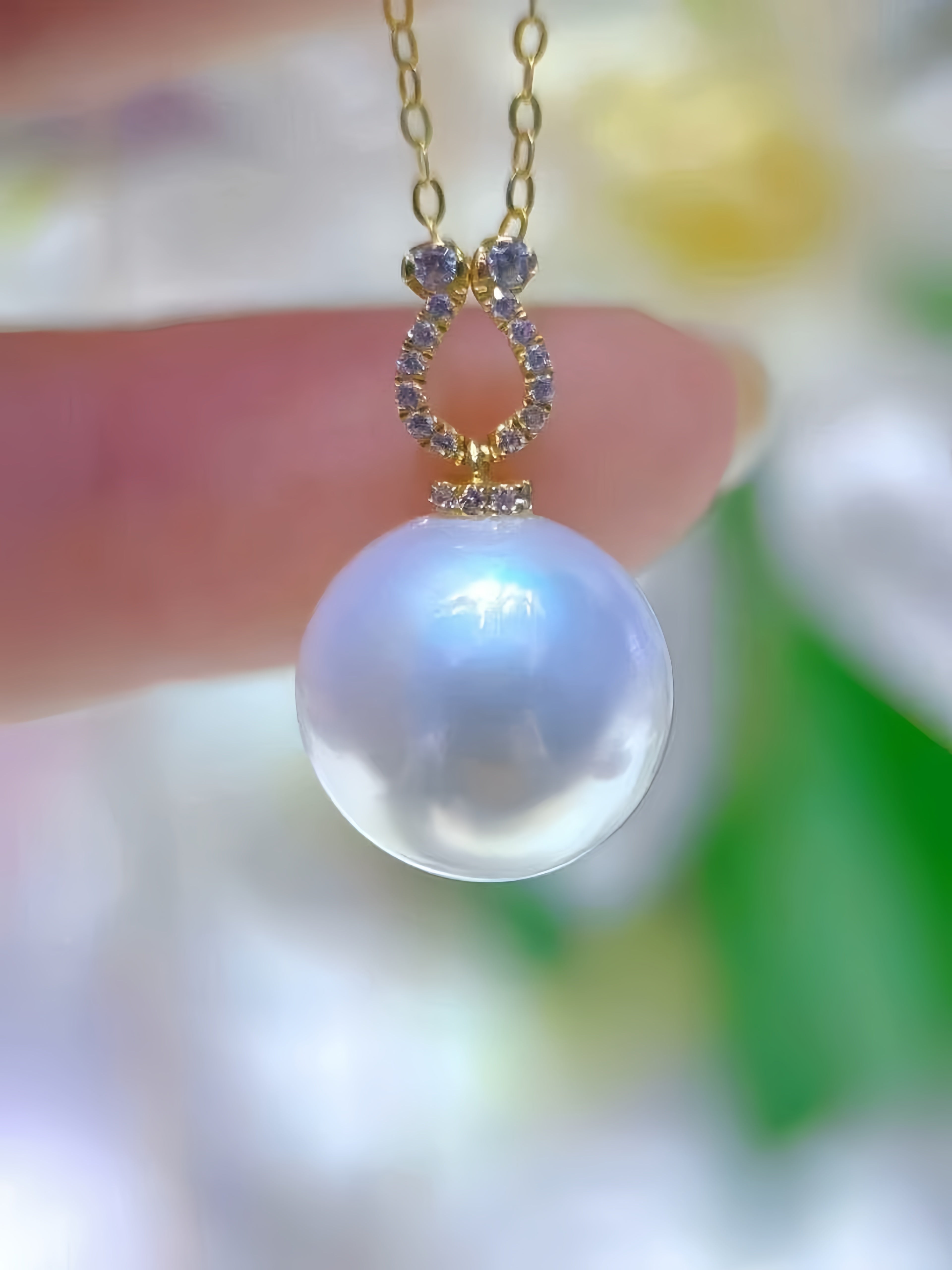 A close-up of a gold pendant necklace with a large, lustrous white pearl and small clear stones, held by a person's fingers against a soft, blurred background.