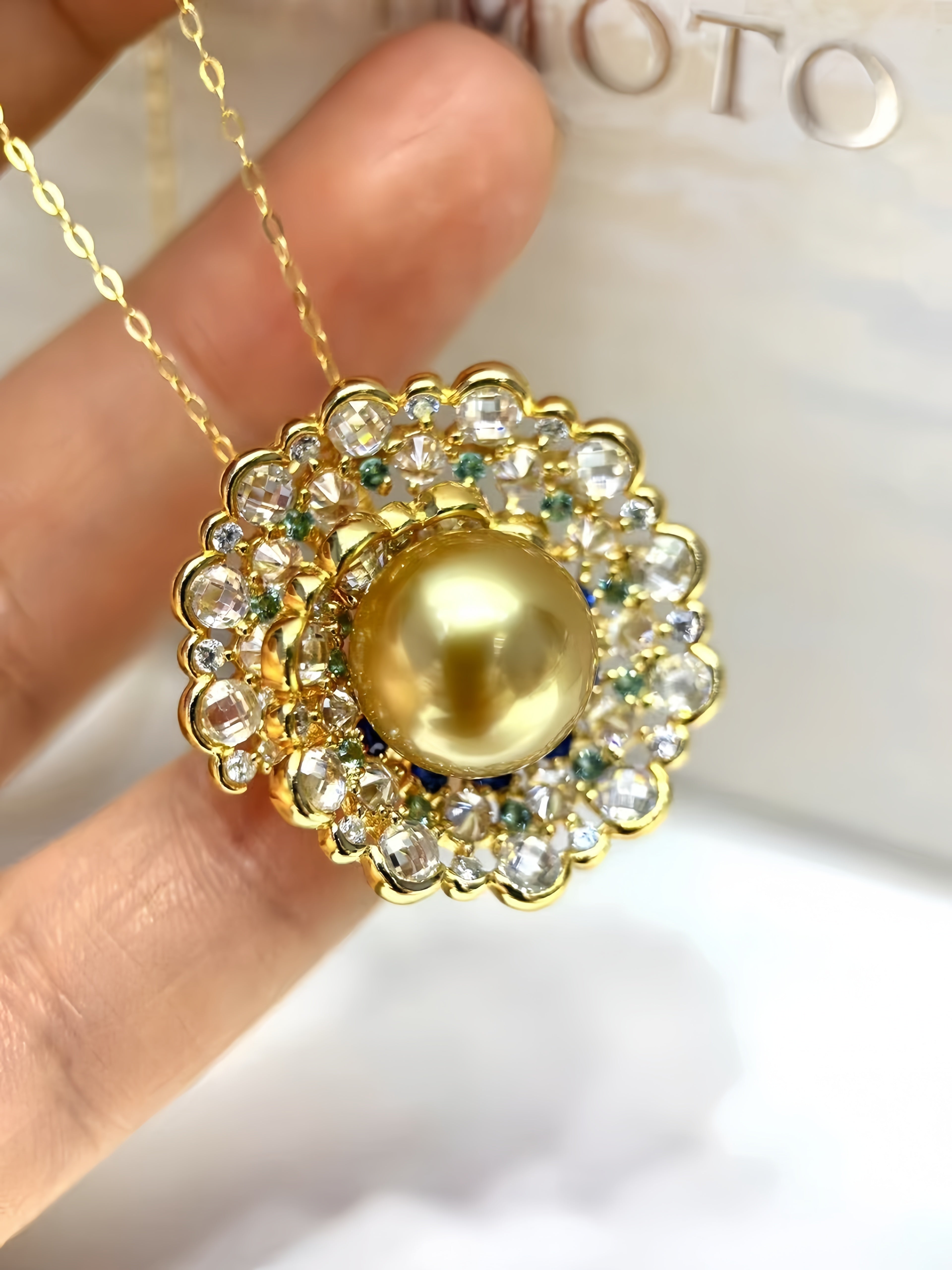 A gold pendant necklace with a large central pearl-like bead surrounded by clear rhinestones and small green accents, held between fingers against a light background.