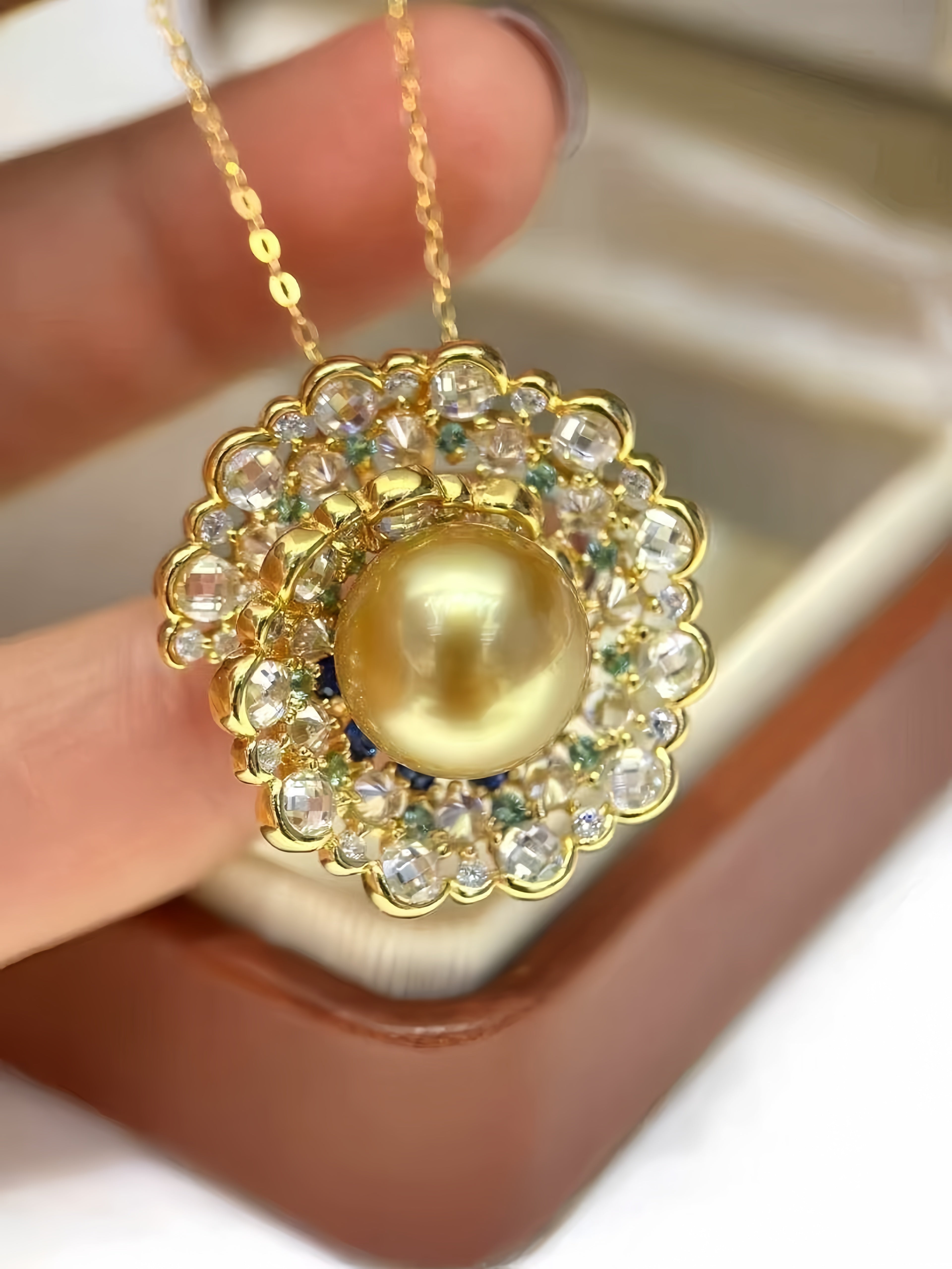 A gold pendant necklace with a large round pearl surrounded by clear rhinestones and small green accents, held by a hand over a brown jewelry box.