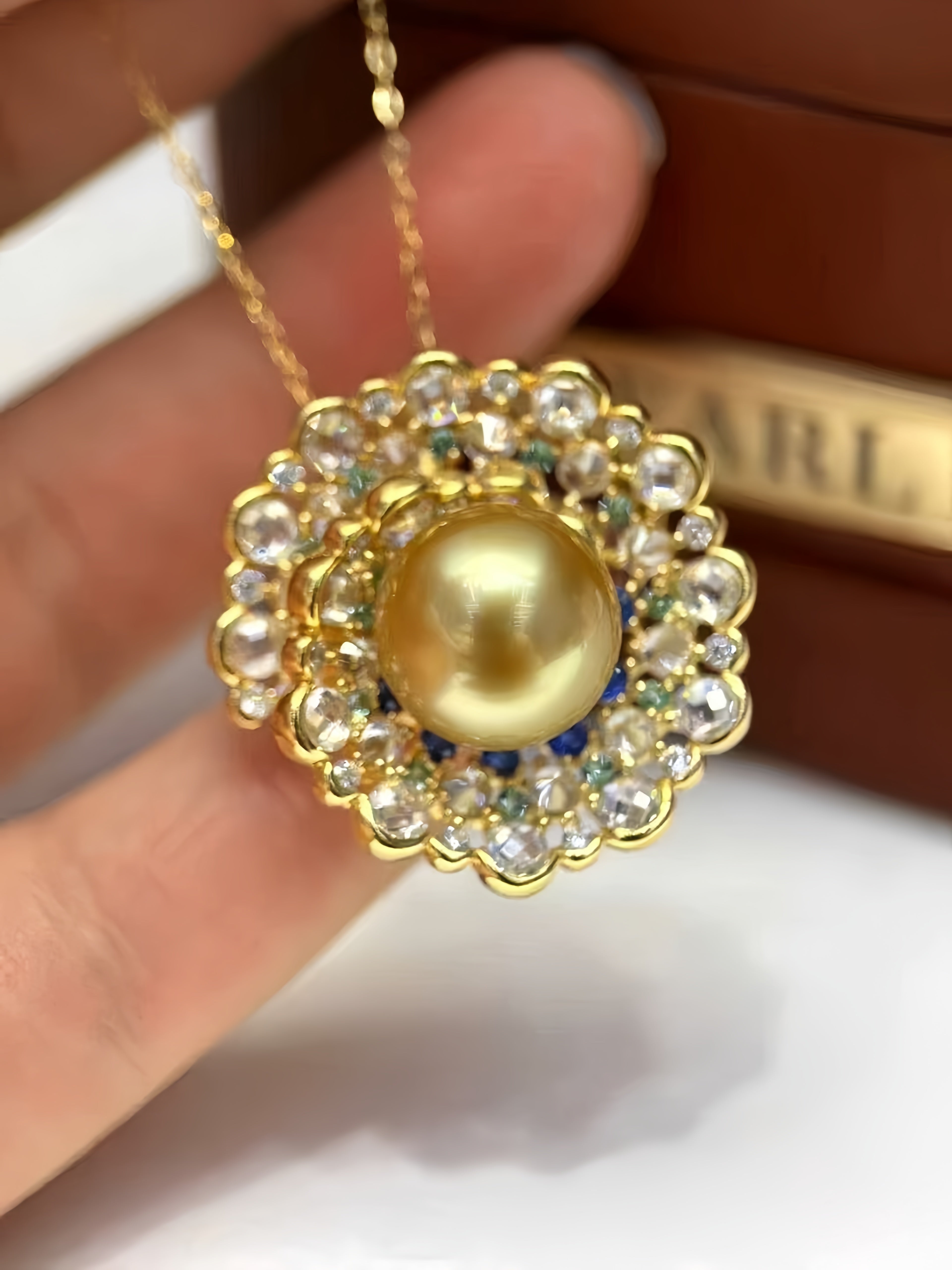 A gold pendant necklace with a large round yellow pearl surrounded by clear rhinestones and small blue and green accents, held by a hand in front of a brown box.