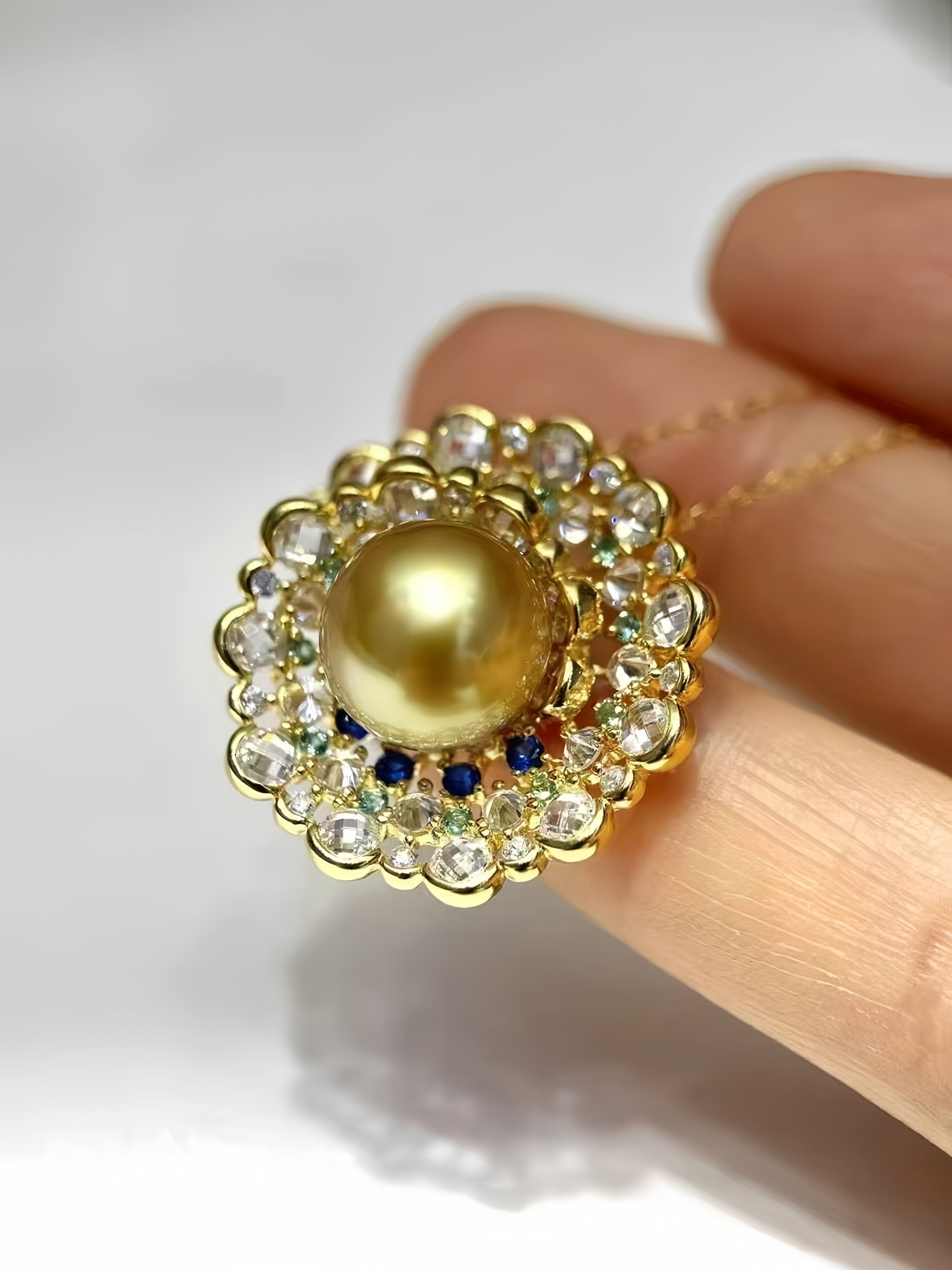 A gold pendant necklace with a large central gold pearl, surrounded by a halo of clear rhinestones and small blue and green accent stones, held between fingers against a white background.