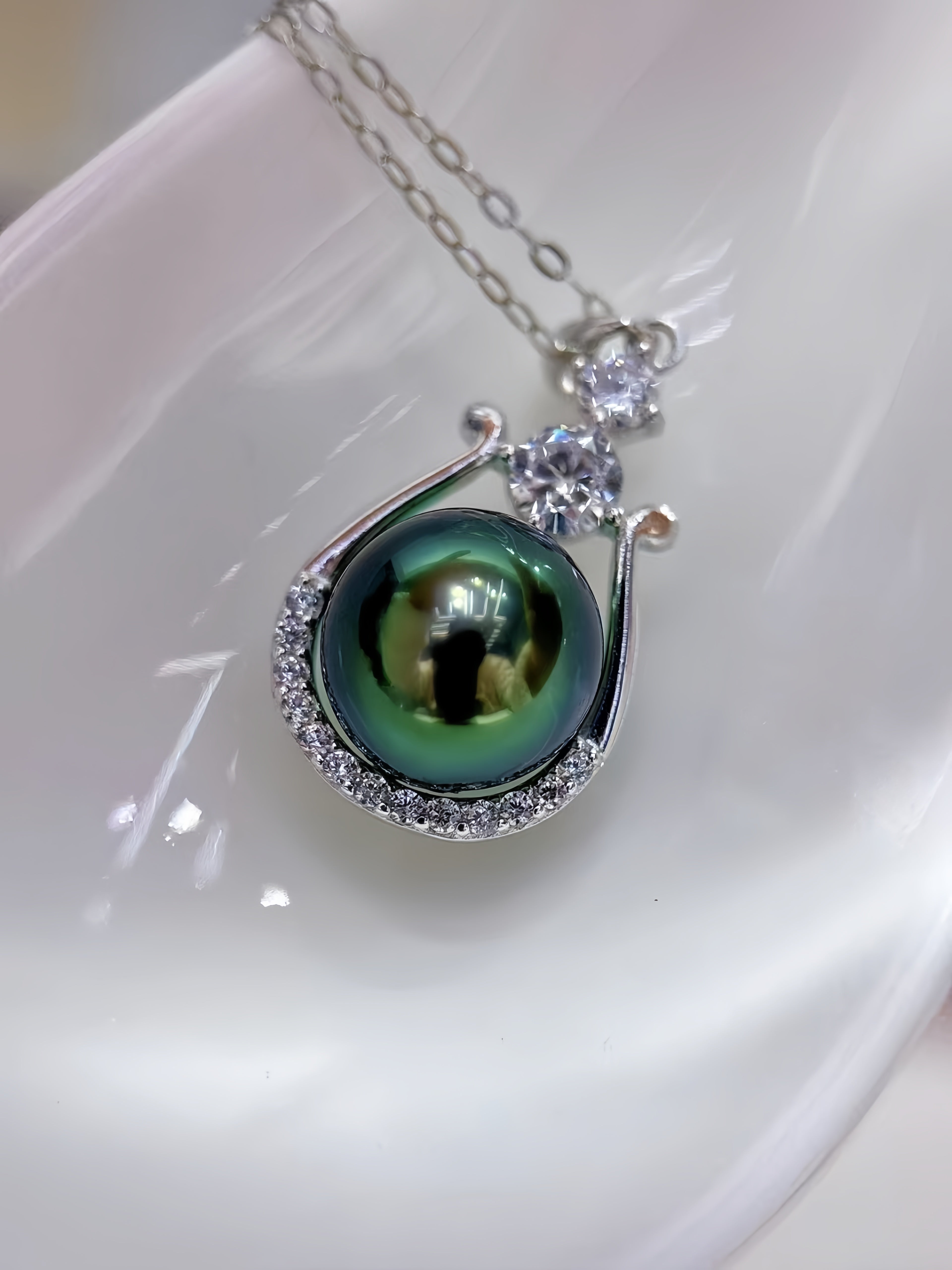 A close-up of a silver pendant necklace featuring a round green pearl surrounded by small clear stones, hanging on a silver chain against a glossy white background.