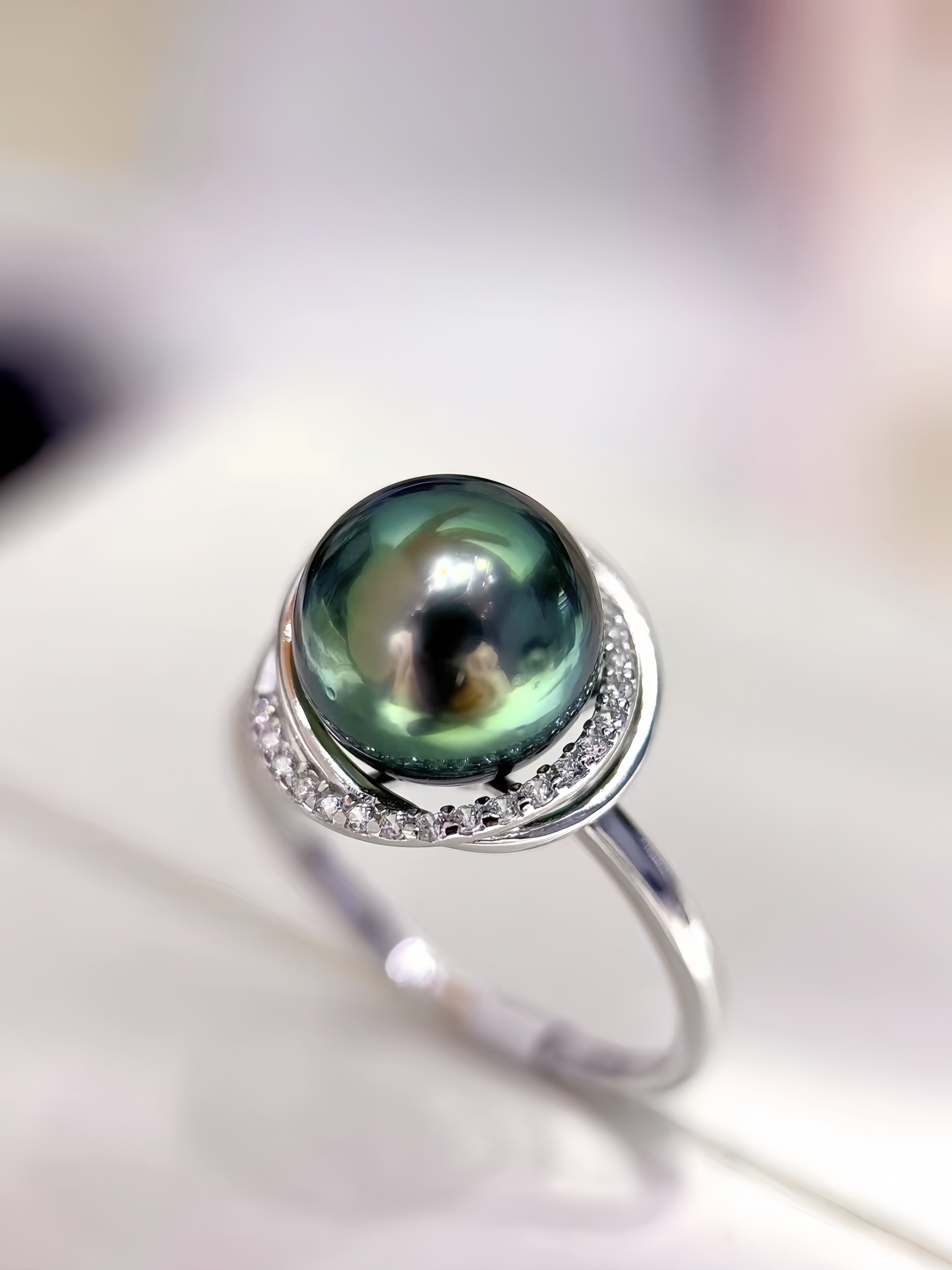 A sterling silver ring featuring a large, round, dark green-black Tahitian pearl accented by small clear stones along the halo, set on a soft-focus light background.