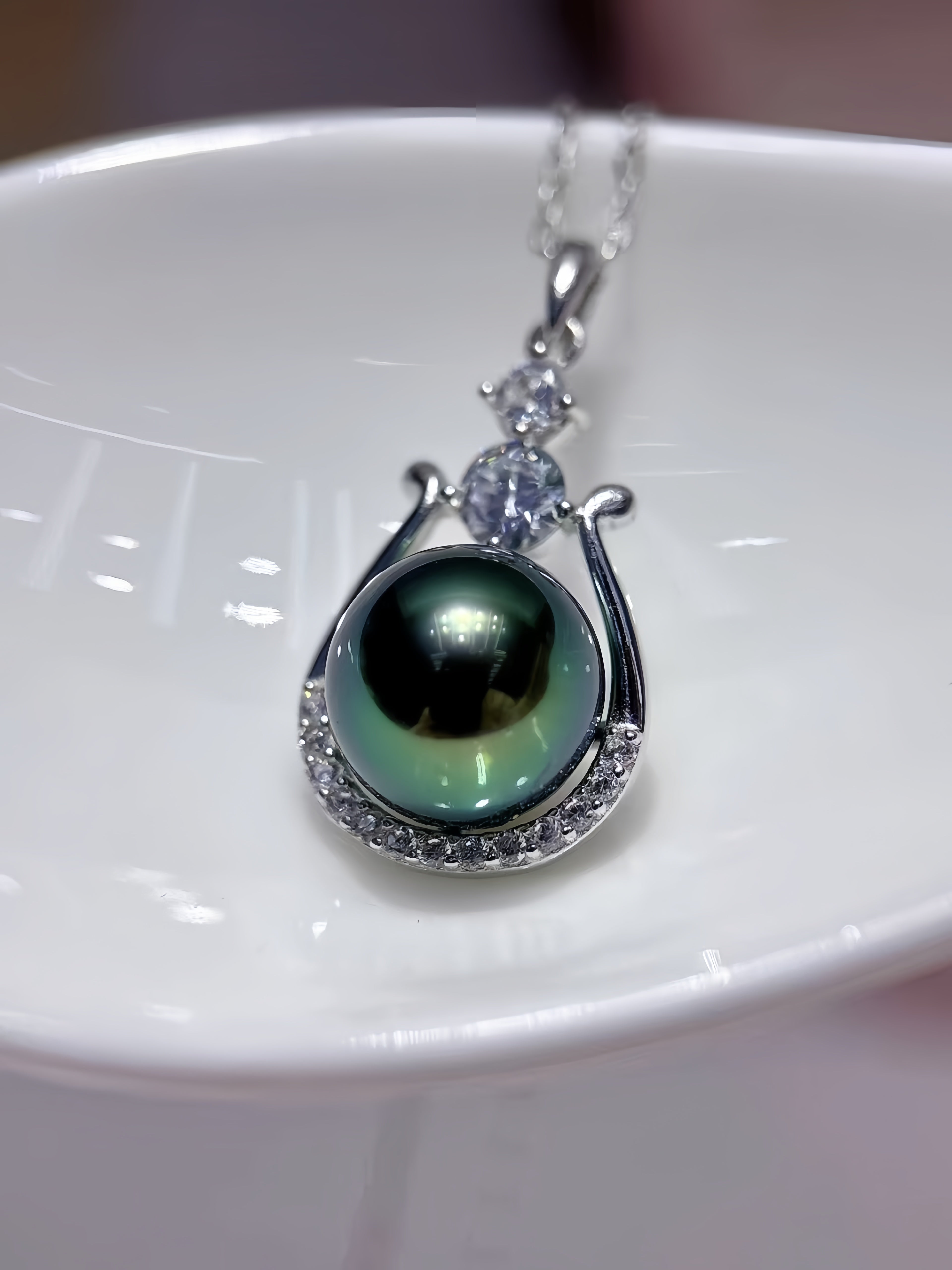 A silver pendant necklace featuring a large, round, dark green iridescent pearl surrounded by small clear stones, resting on a white ceramic dish.