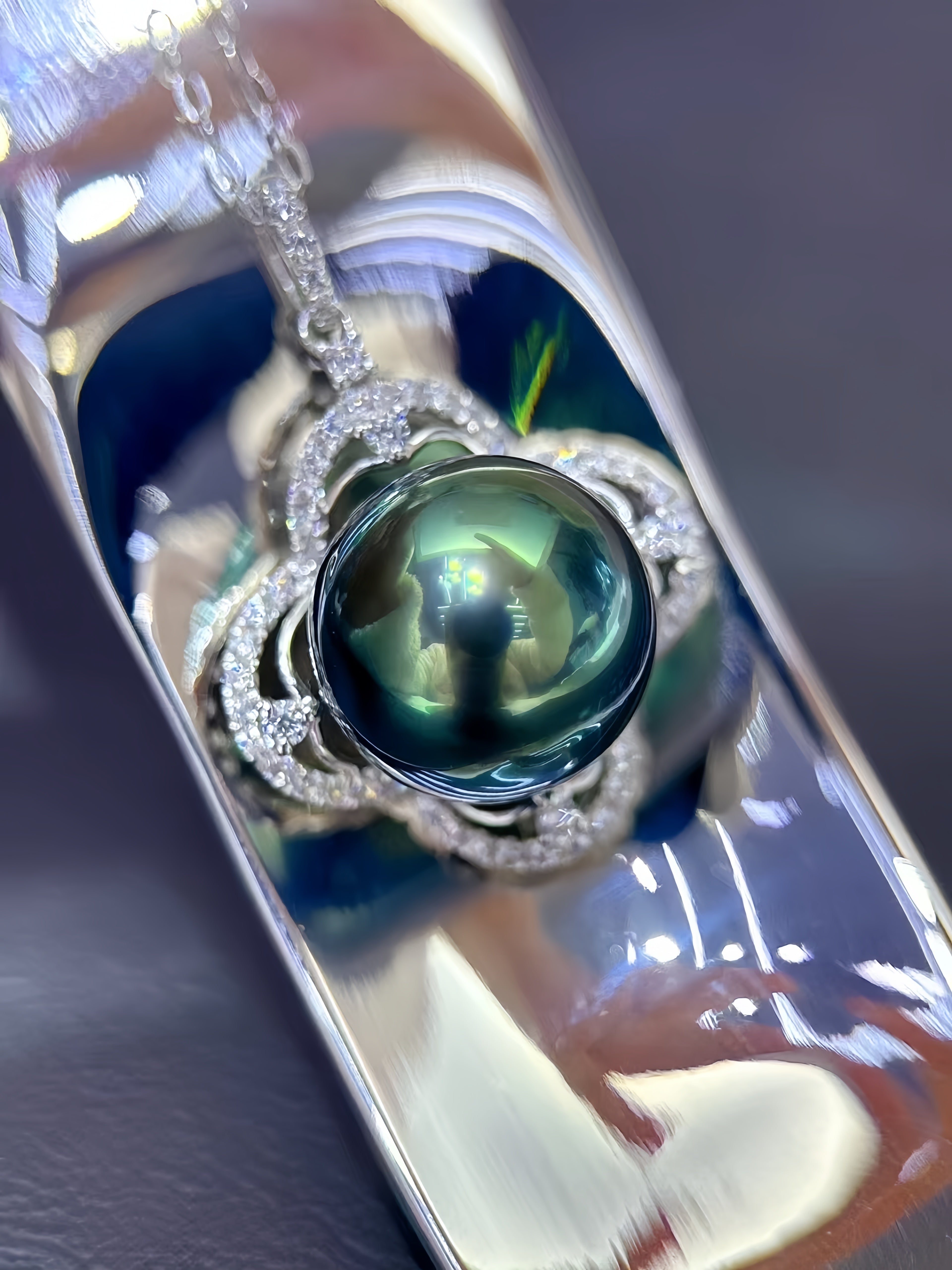 A close-up of a silver pendant necklace featuring a large, round, dark green teardrop-shaped pearl surrounded by a halo of small clear stones, resting on a highly reflective silver surface.
