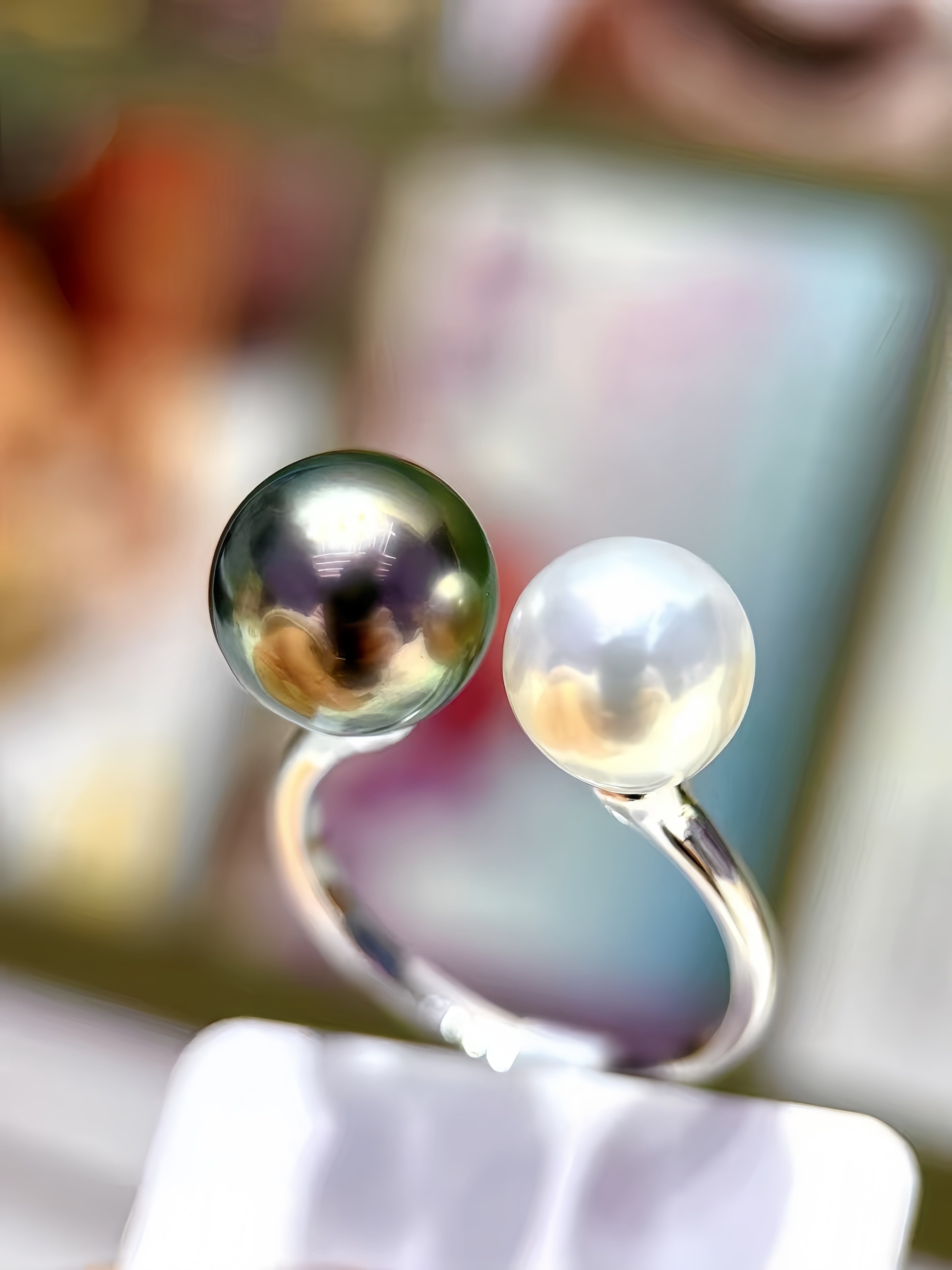 A close-up of a silver-toned ring with two round pearls, one dark green-black and one white, displayed on a stand against a blurred background.