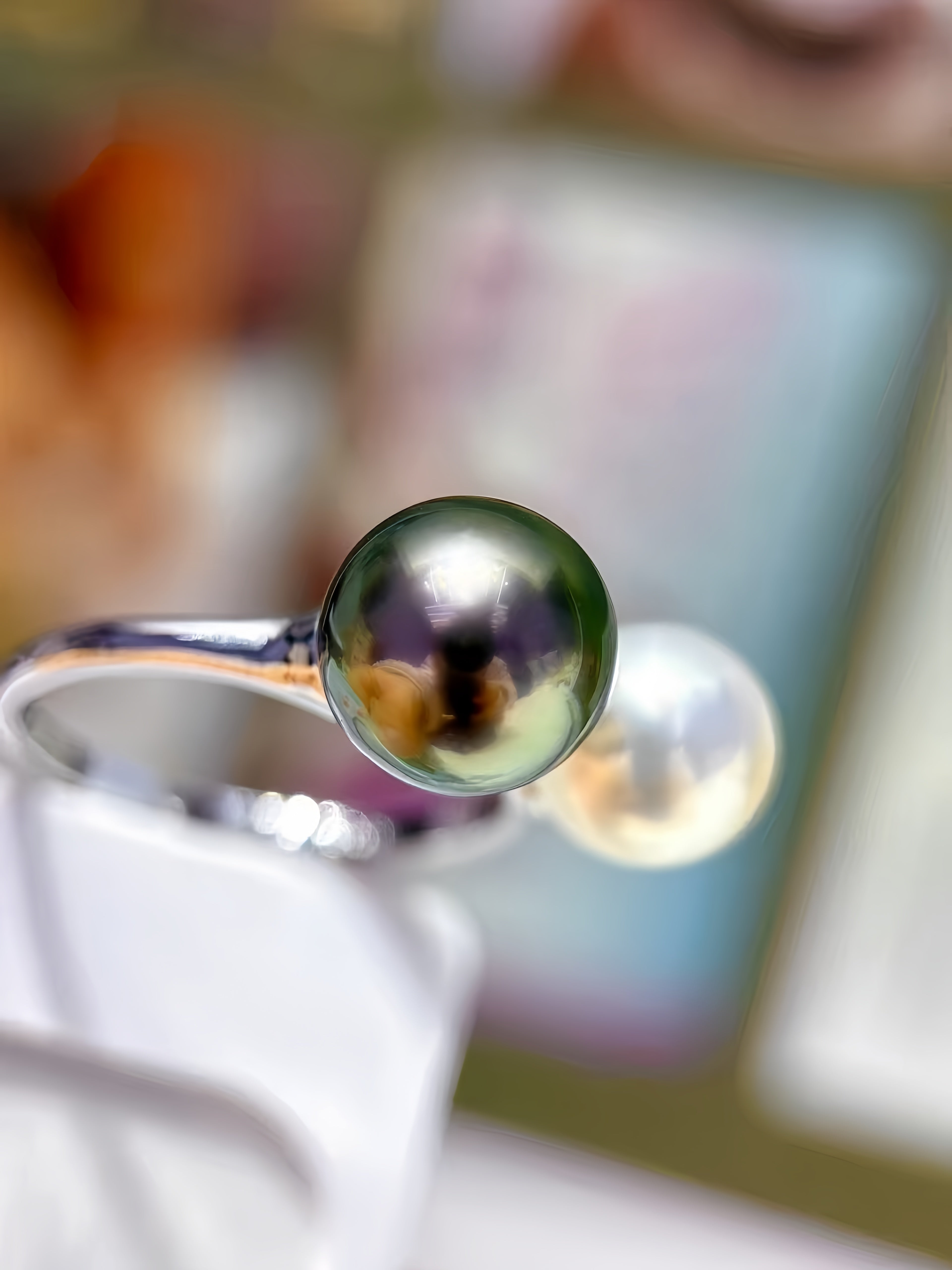 A close-up of a polished silver ring holding a large, dark green-black freshwater pearl on a white display stand, with a second pearl in the background.