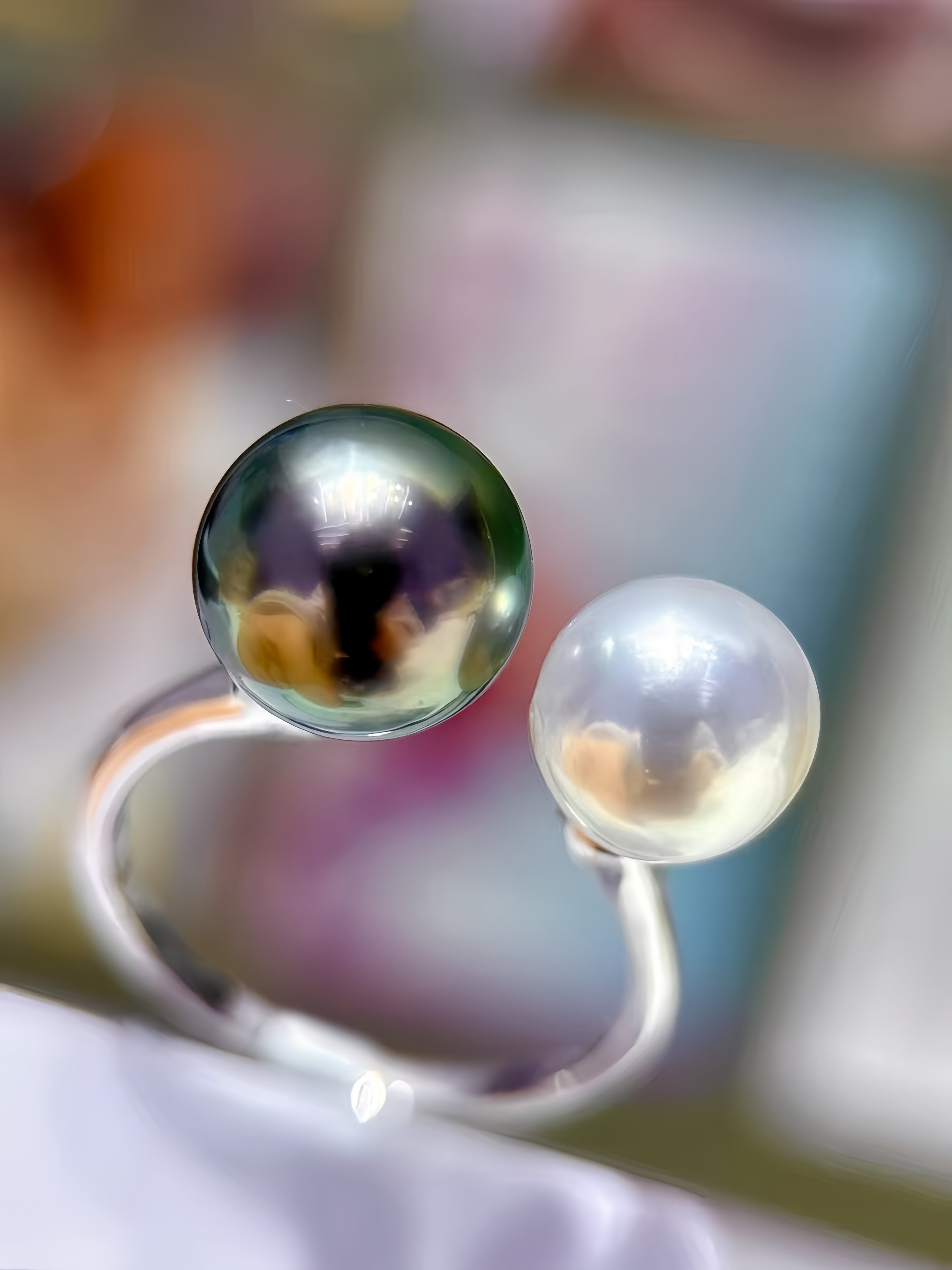 A silver-toned ring featuring two spherical pearl-like beads, one dark iridescent and one pale milky white, photographed close up against a softly blurred background.