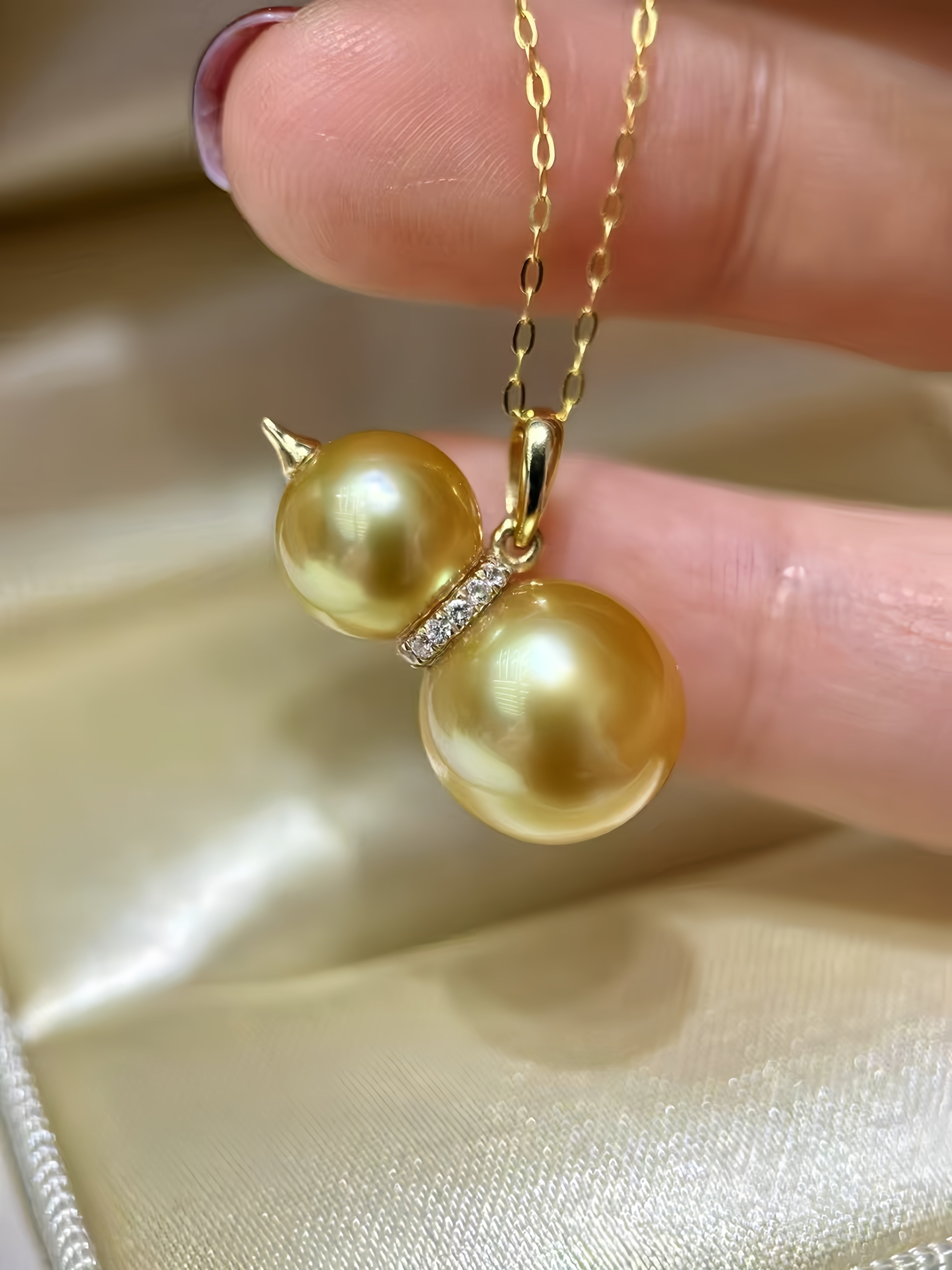 A gold pendant necklace with two large gold freshwater pearls connected by a small diamond-accented spacer, held by fingers over a champagne-colored satin surface.