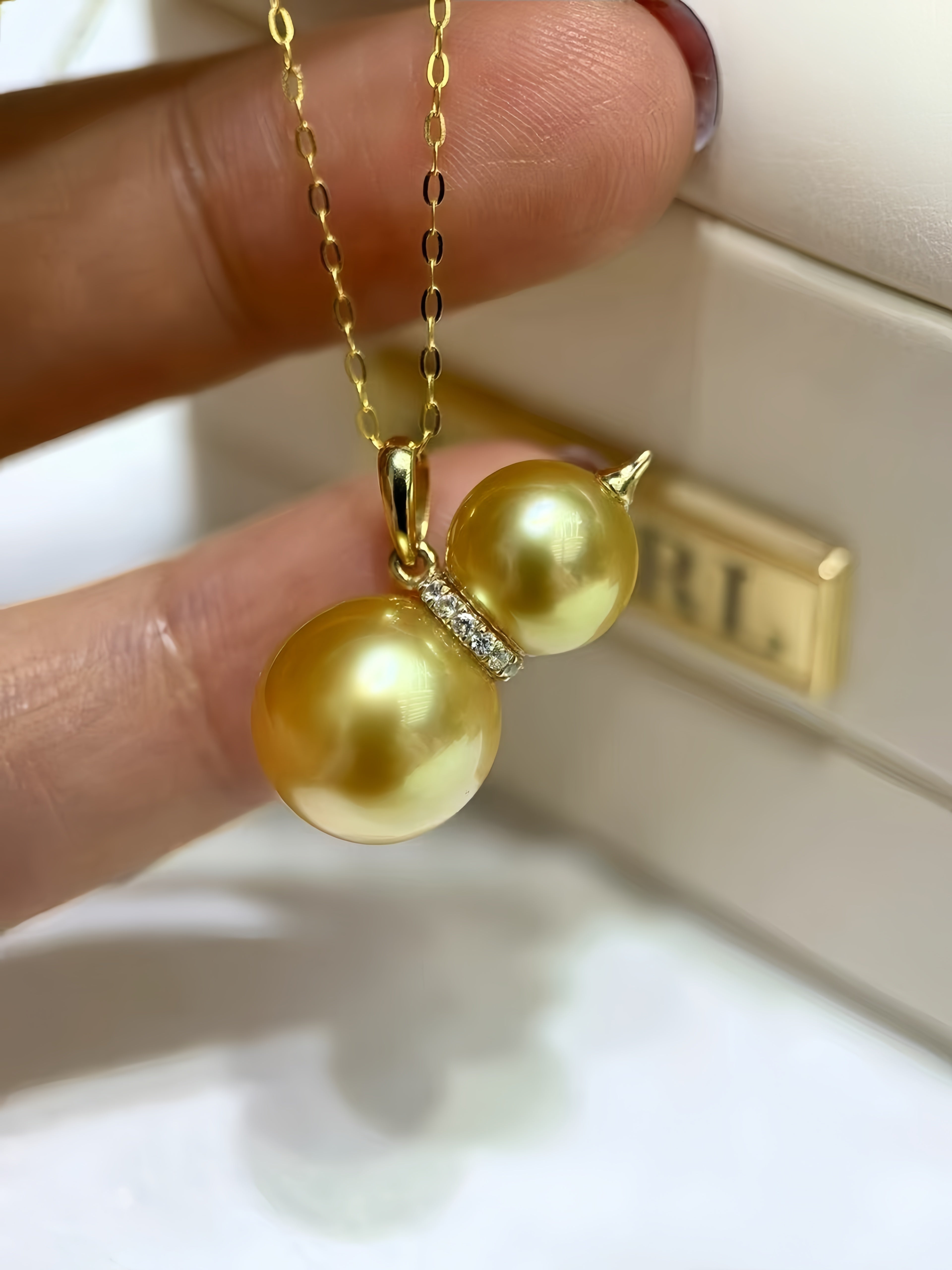 A gold pendant necklace with two large round gold pearls connected by a small clear stone accent, held between fingers in front of a cream jewelry box.