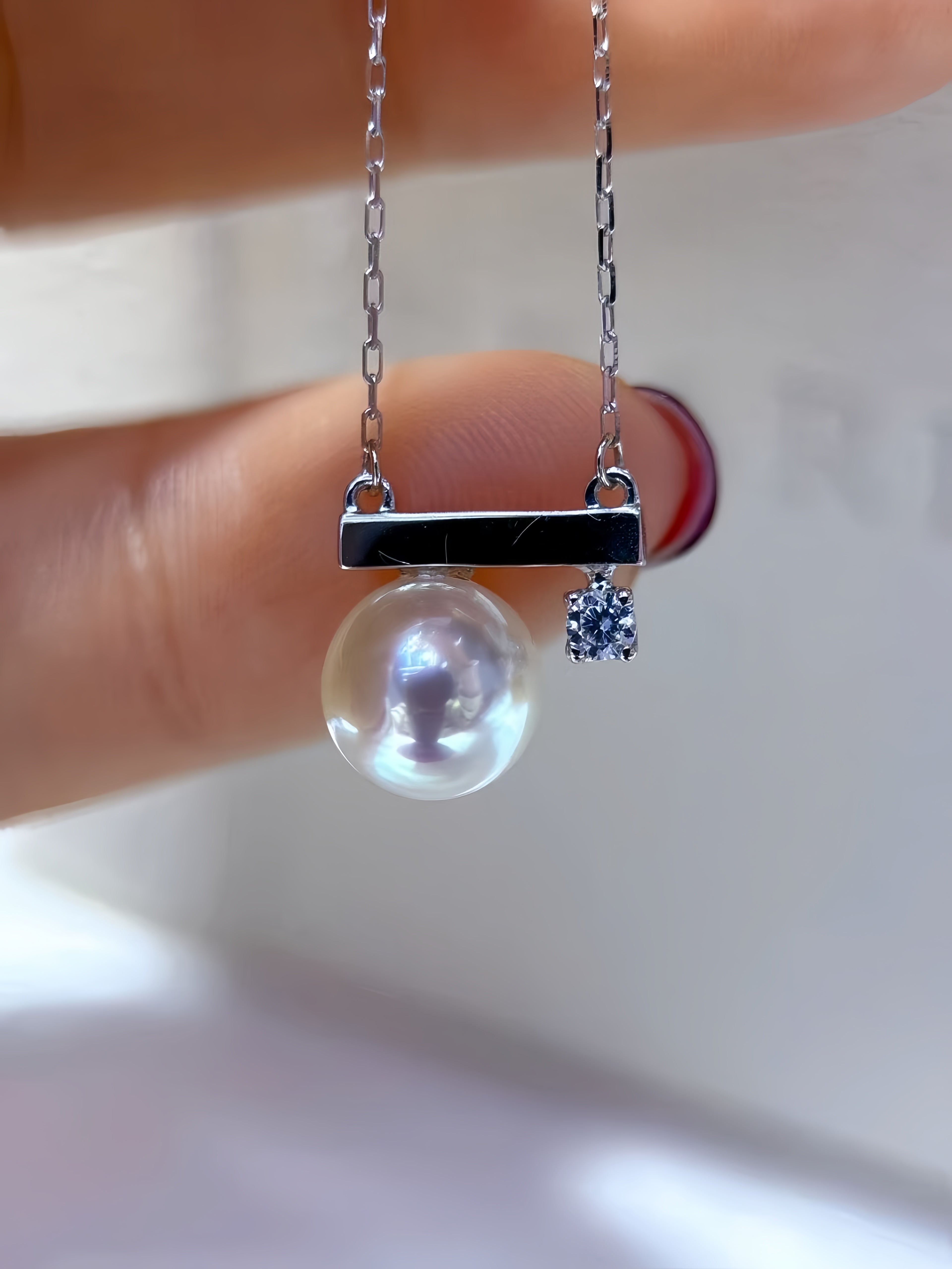 A close-up of a silver necklace held between fingers, featuring a round white pearl pendant, a horizontal silver bar, and a small dangling blue gemstone.
