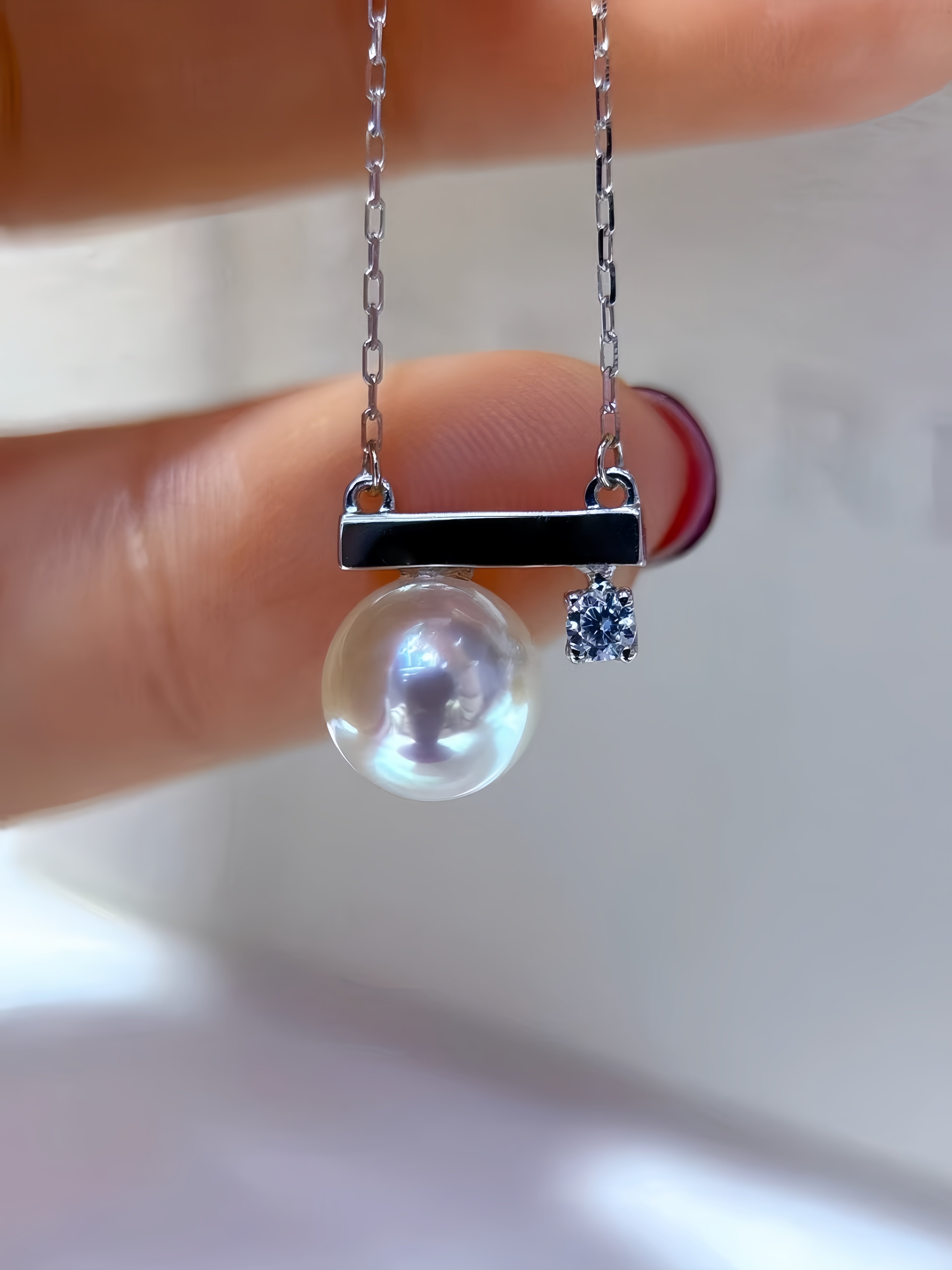 A close-up of a delicate necklace held between fingers, featuring a silver bar pendant with a large round white pearl and a small sparkling blue gemstone charm, set against a soft white background.