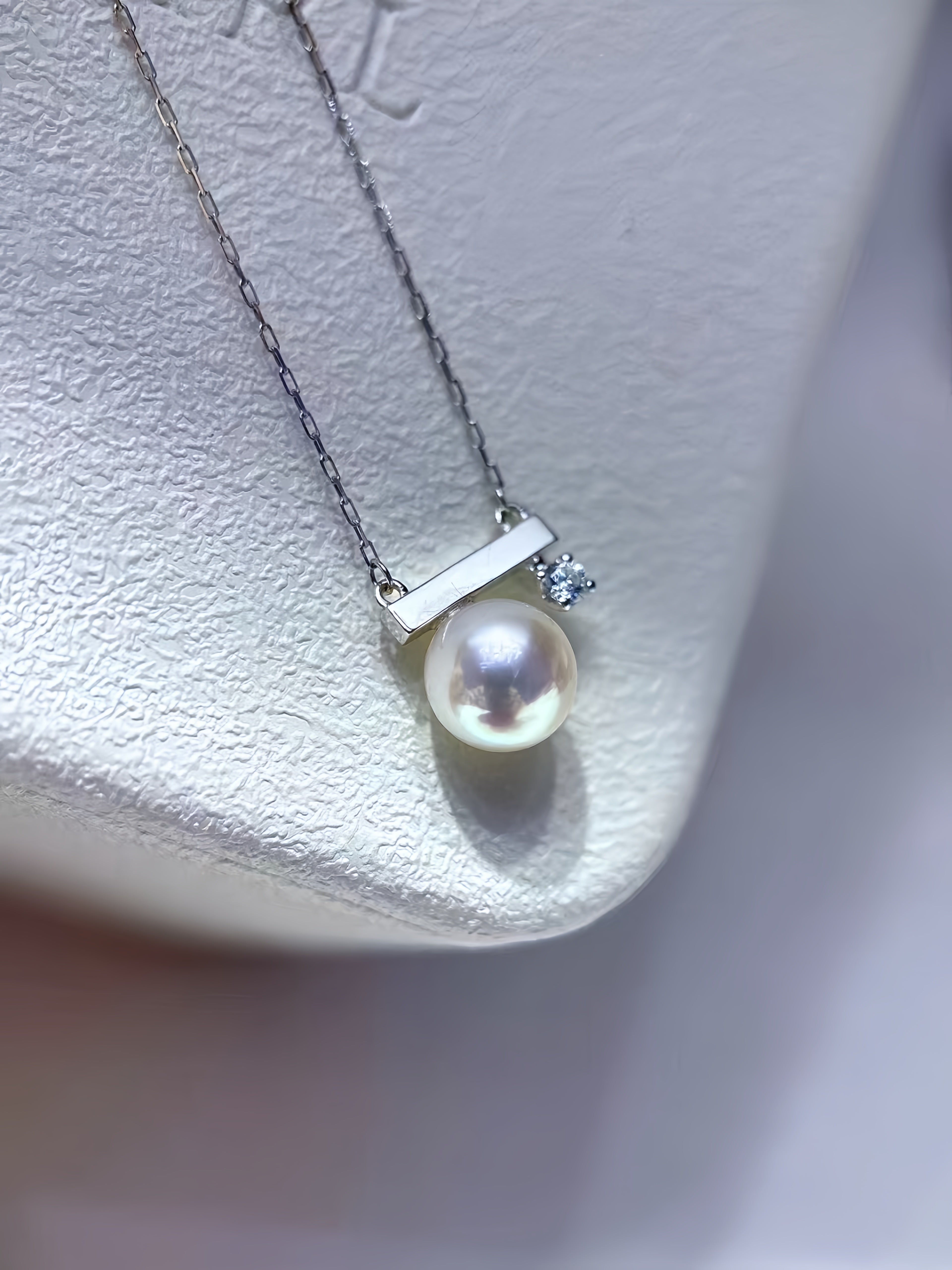 A delicate silver necklace featuring a round white pearl pendant and a small clear accent stone, displayed on a textured white jewelry stand.
