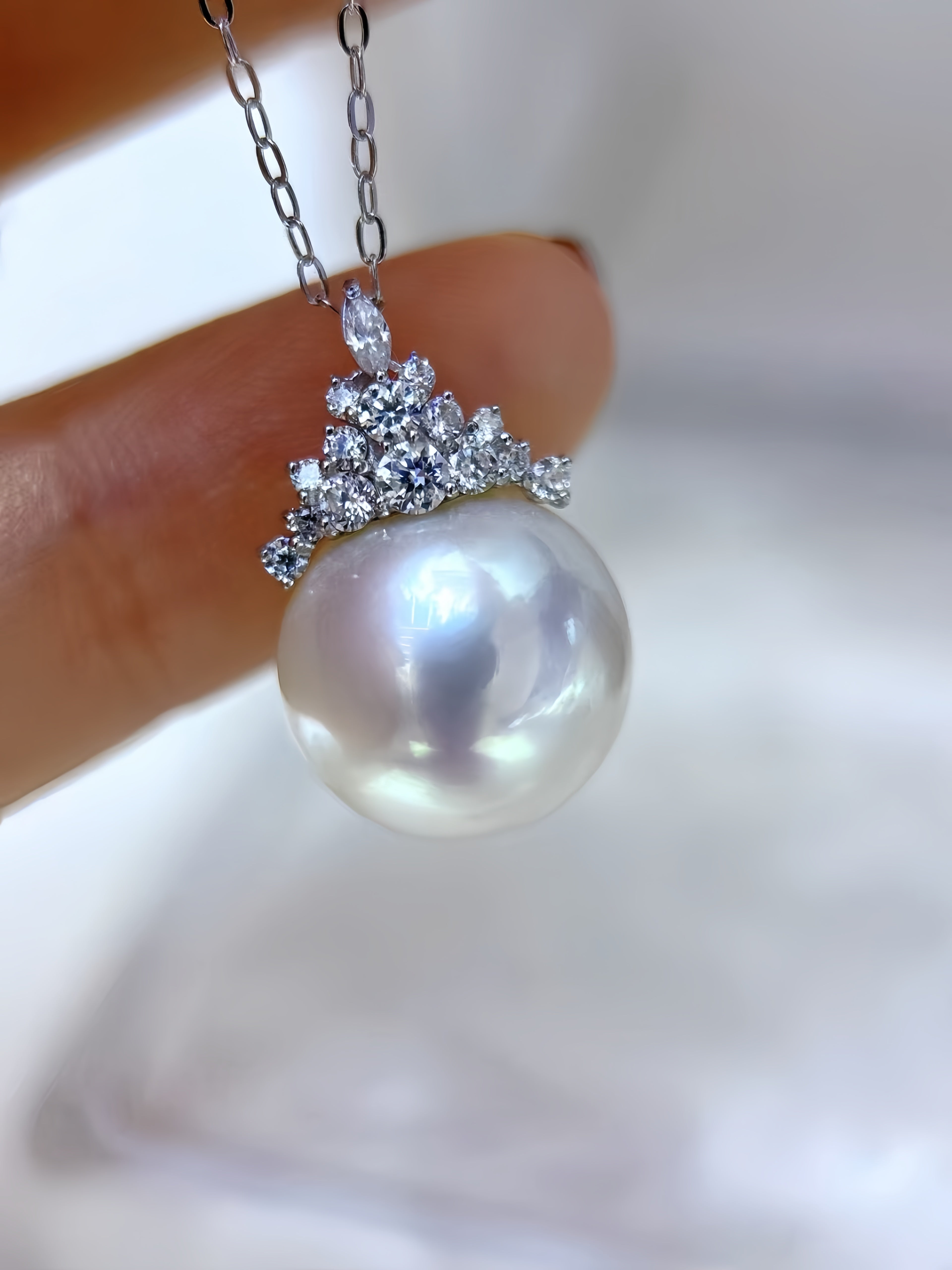 A close-up of a pearl pendant necklace held between fingers. The pendant features a large, lustrous white pearl suspended below a silver-toned crown-like setting adorned with small clear stones, on a delicate chain.