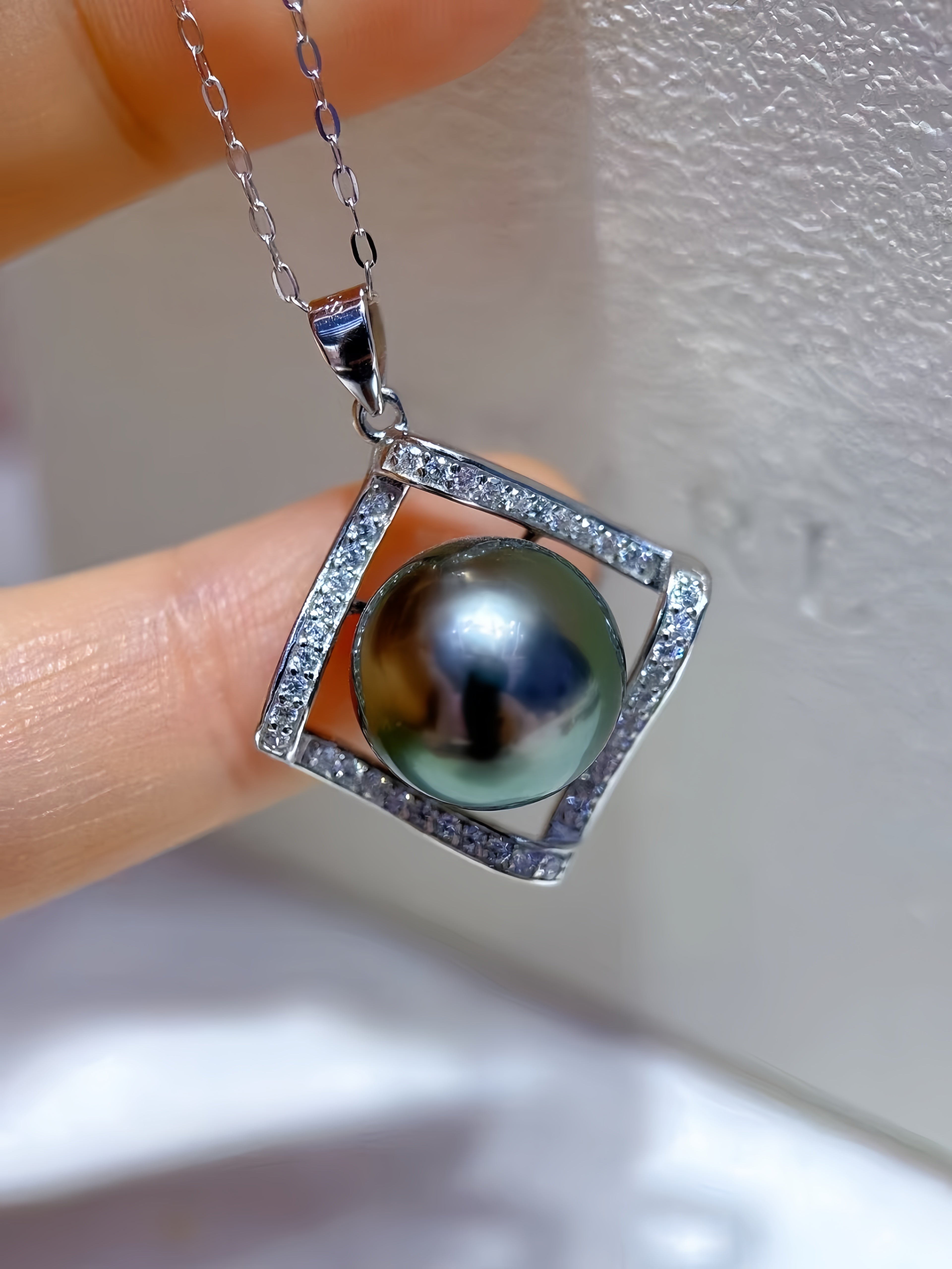 A close-up of a hand holding a silver pendant necklace featuring a round iridescent green pearl surrounded by a square halo of small clear stones.