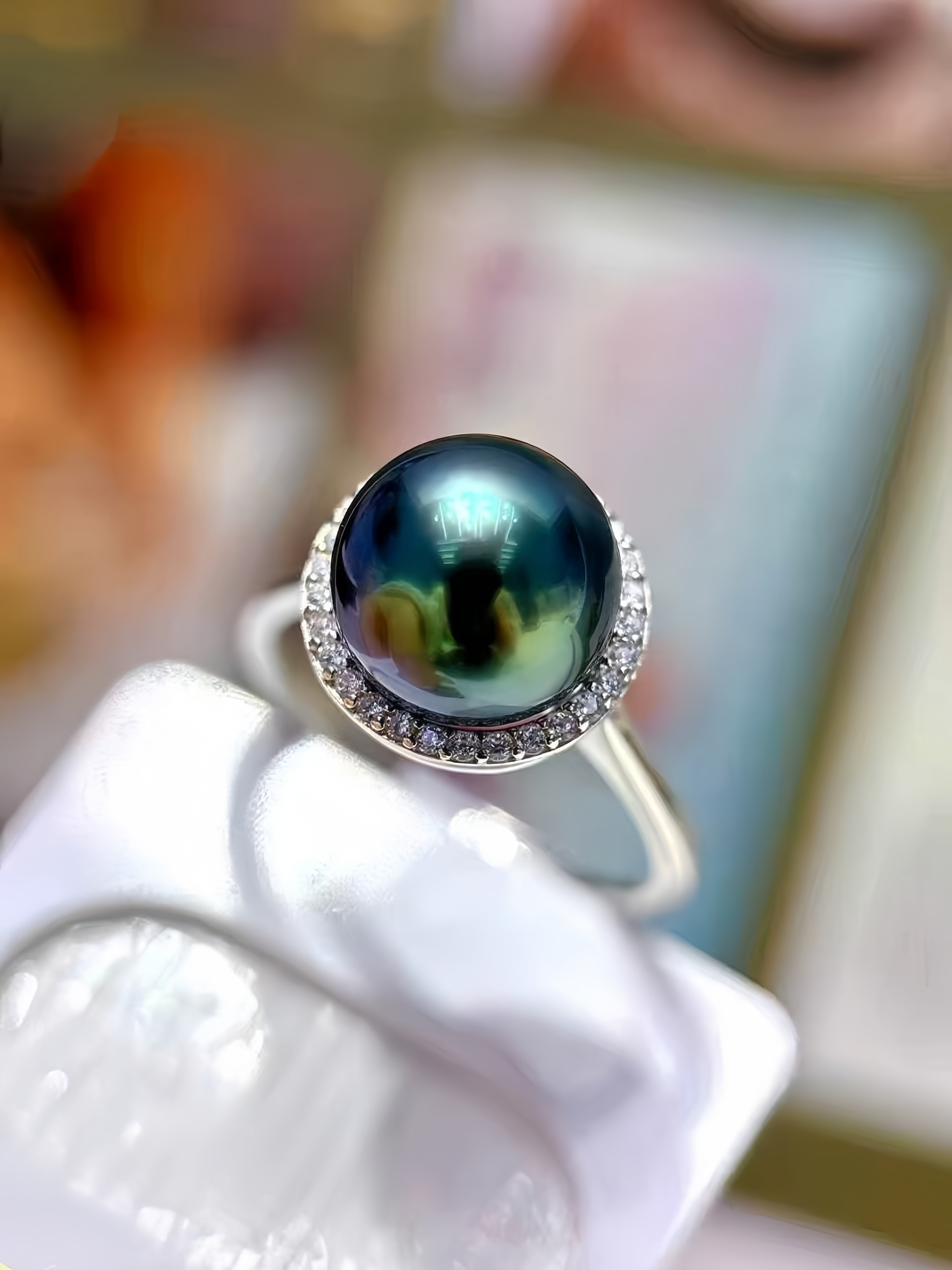 A close-up of a ring featuring a round iridescent dark green pearl surrounded by a halo of small clear stones, displayed on a white stand.