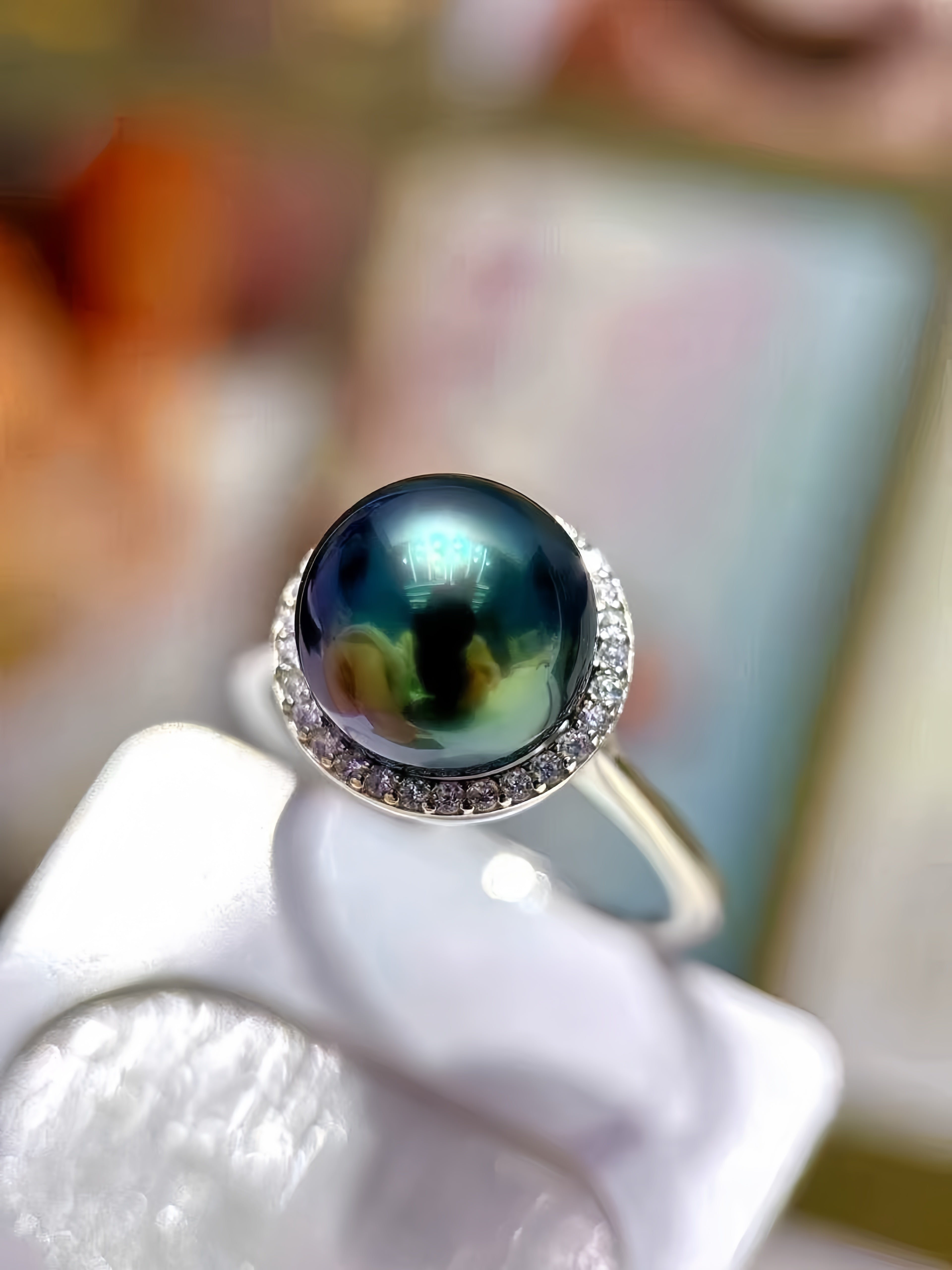 A close-up of a ring featuring a large round iridescent dark pearl with a halo of small clear stones, displayed on a white stand.