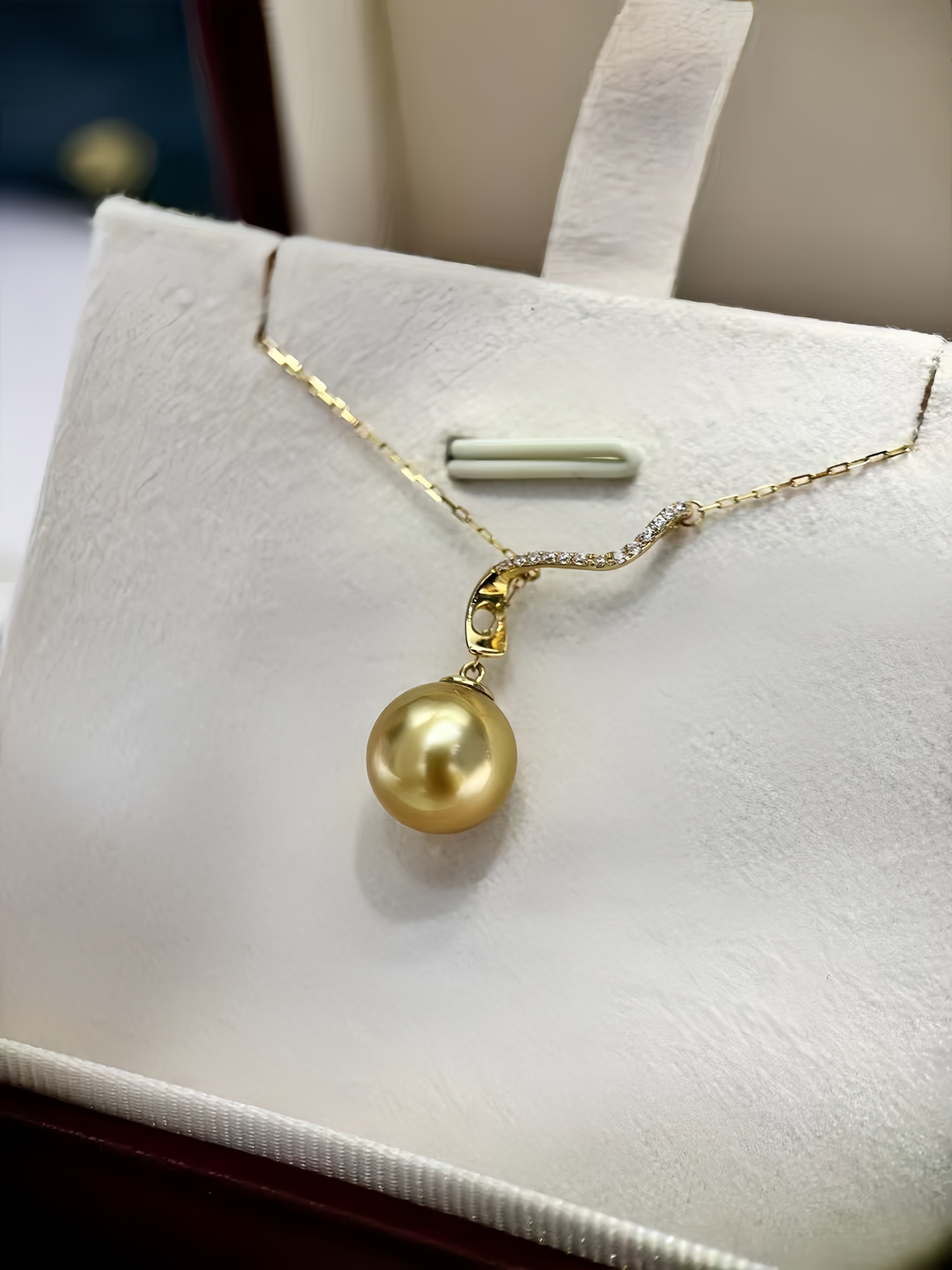 A gold pendant necklace with a single round freshwater pearl charm, displayed on a white jewelry stand inside a box.