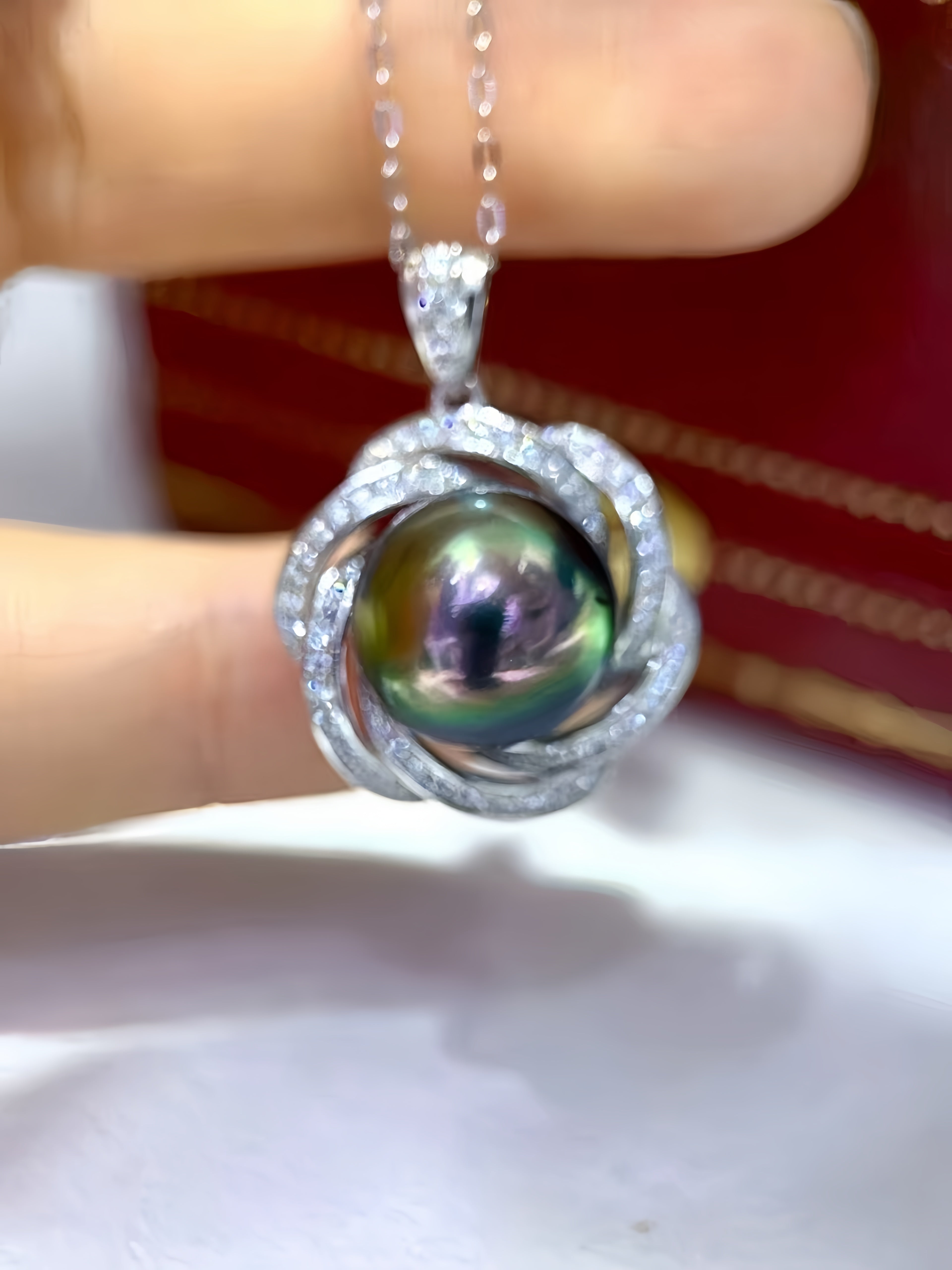 A close-up of a hand holding a silver-toned pendant necklace featuring a round iridescent green pearl surrounded by small clear stones, with a red box blurred in the background.
