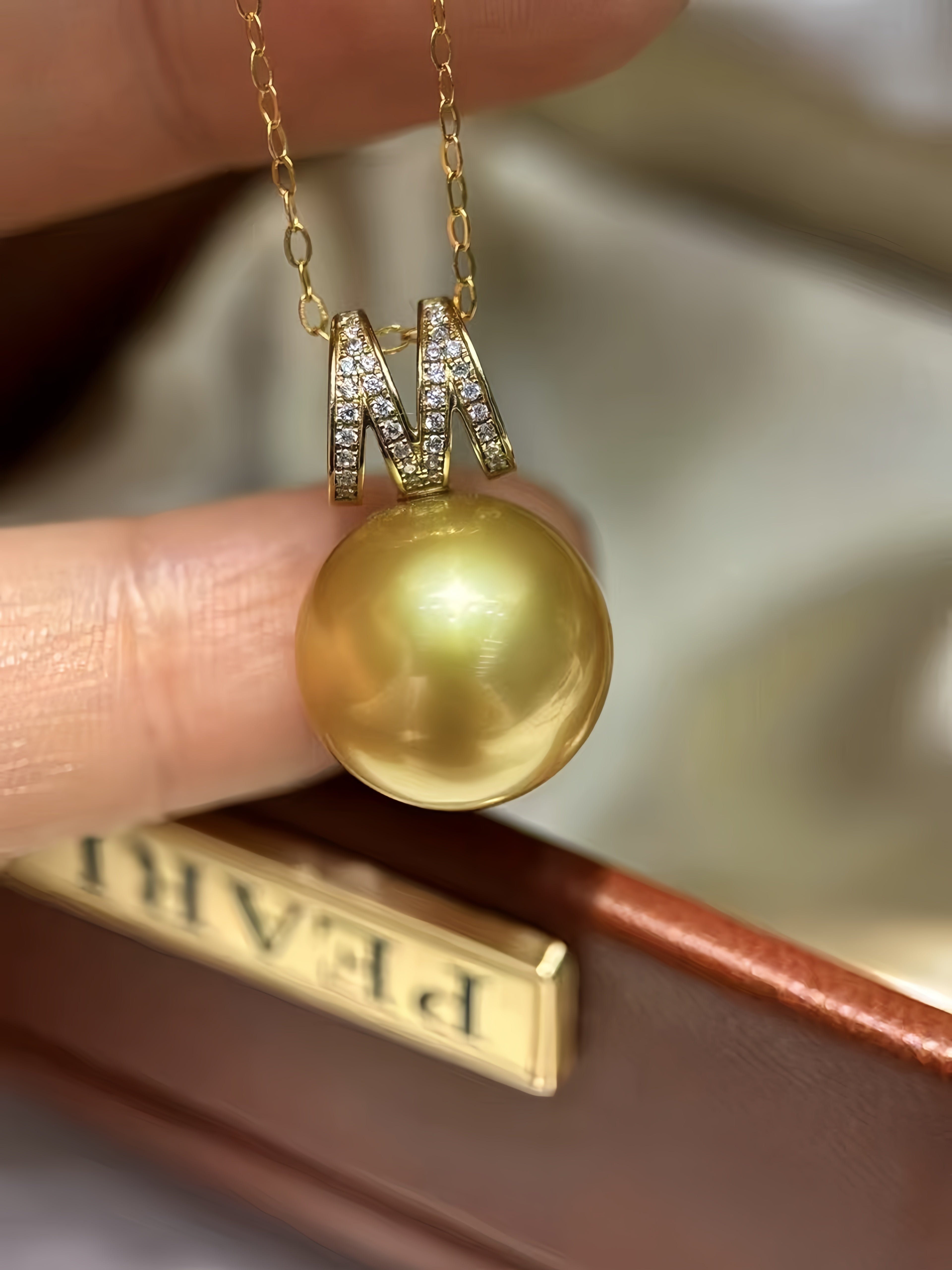 A gold pendant necklace featuring a large, round, lustrous gold pearl suspended from a gold chain with a decorative letter M bail set with small clear stones, held by fingers in front of a brown leather box.