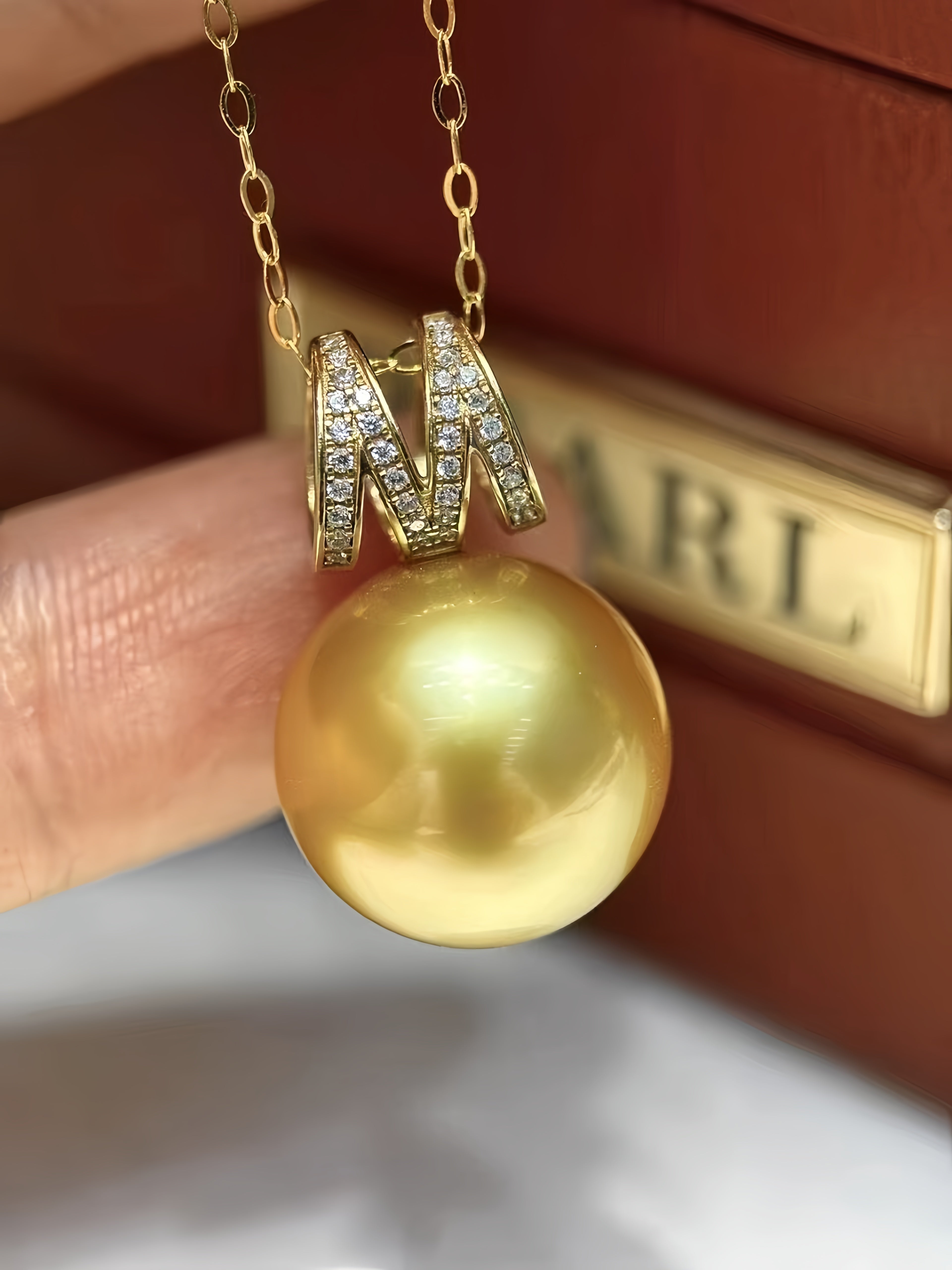 A gold pendant necklace featuring a large, round, lustrous gold pearl and a decorative gold 'M' setting with pavé-set clear stones, held by a hand in front of a brown box with a gold 'MARL' logo.
