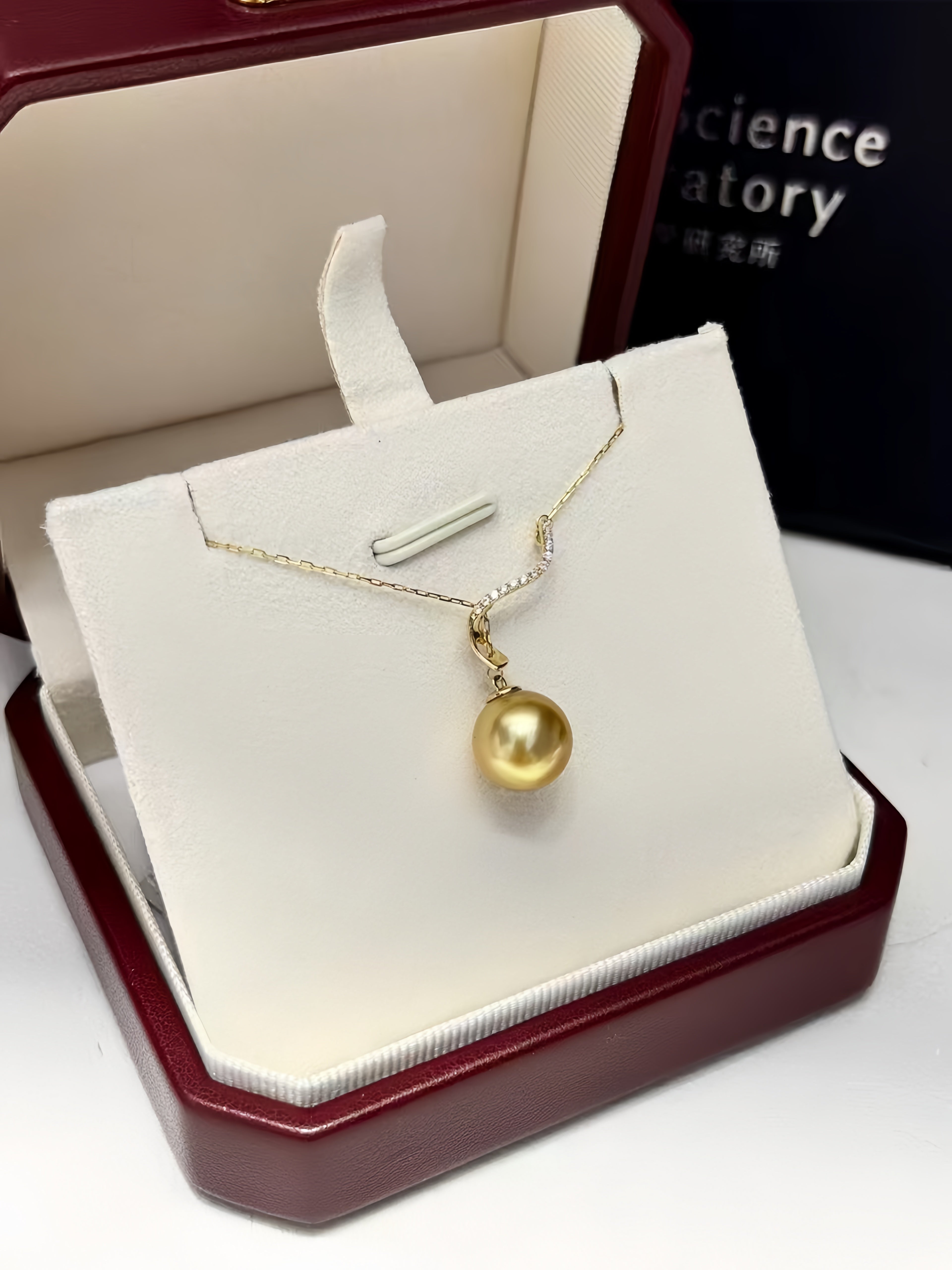 A gold pendant necklace with a single round pearl-like drop, displayed on a white jewelry stand inside an open dark red presentation box. A black box in the background shows the brand name 'science factory'.