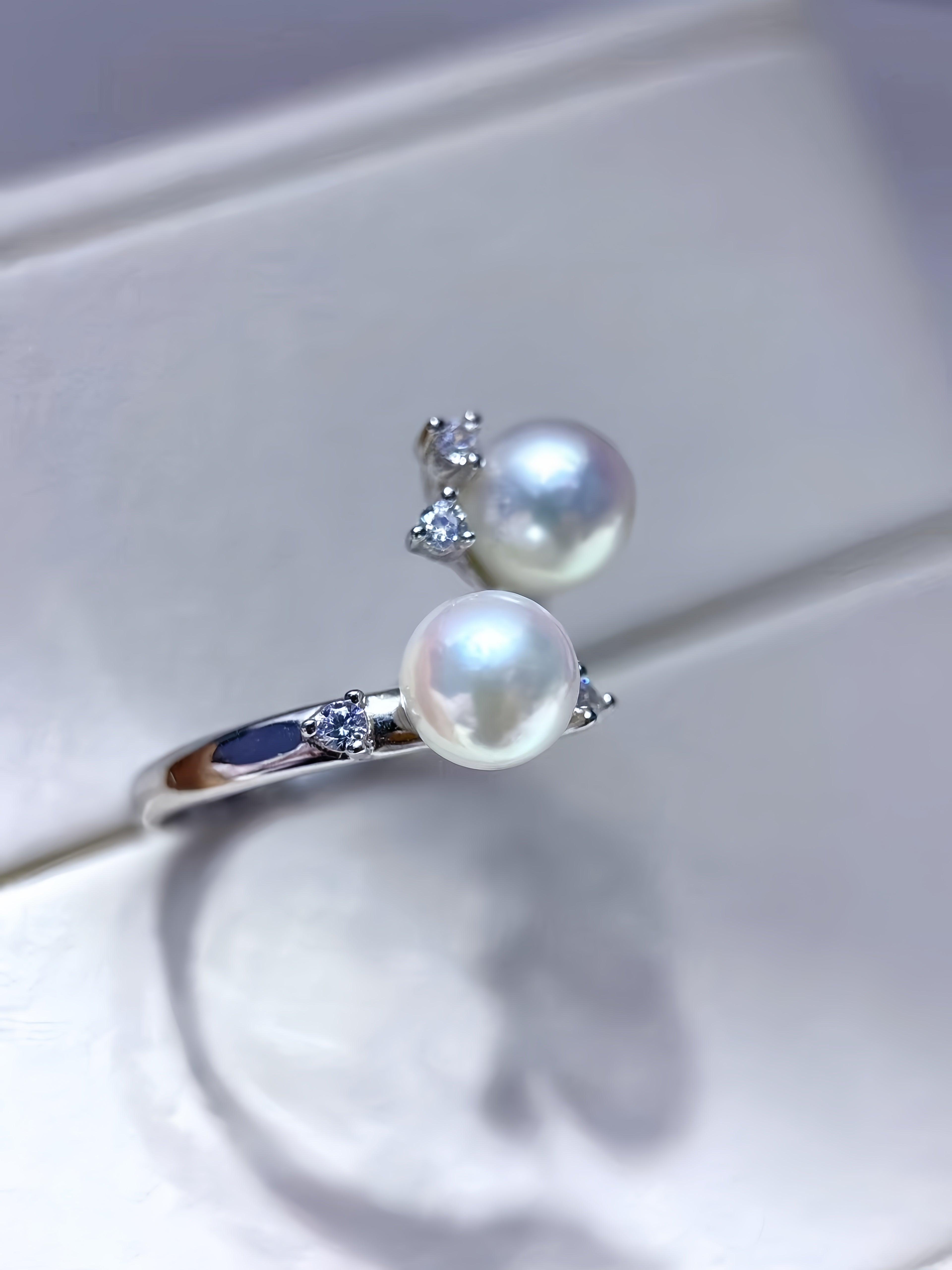 A silver ring featuring two large white pearls and small clear accent stones, displayed on a white ring box.