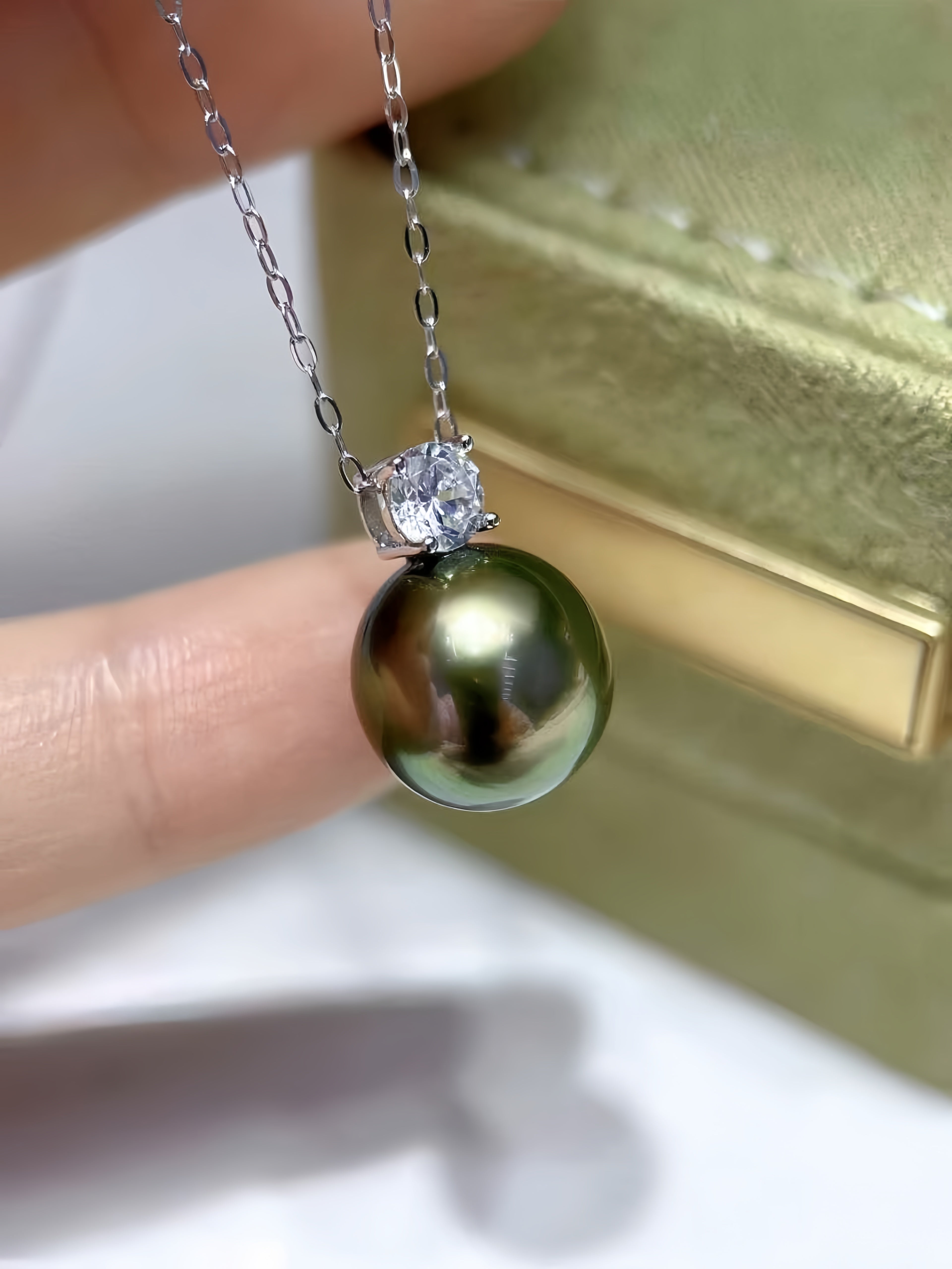 A close-up of a necklace with a round dark green pearl pendant and a clear stone setting on a silver chain, held between fingers with a gold jewelry box in the background.