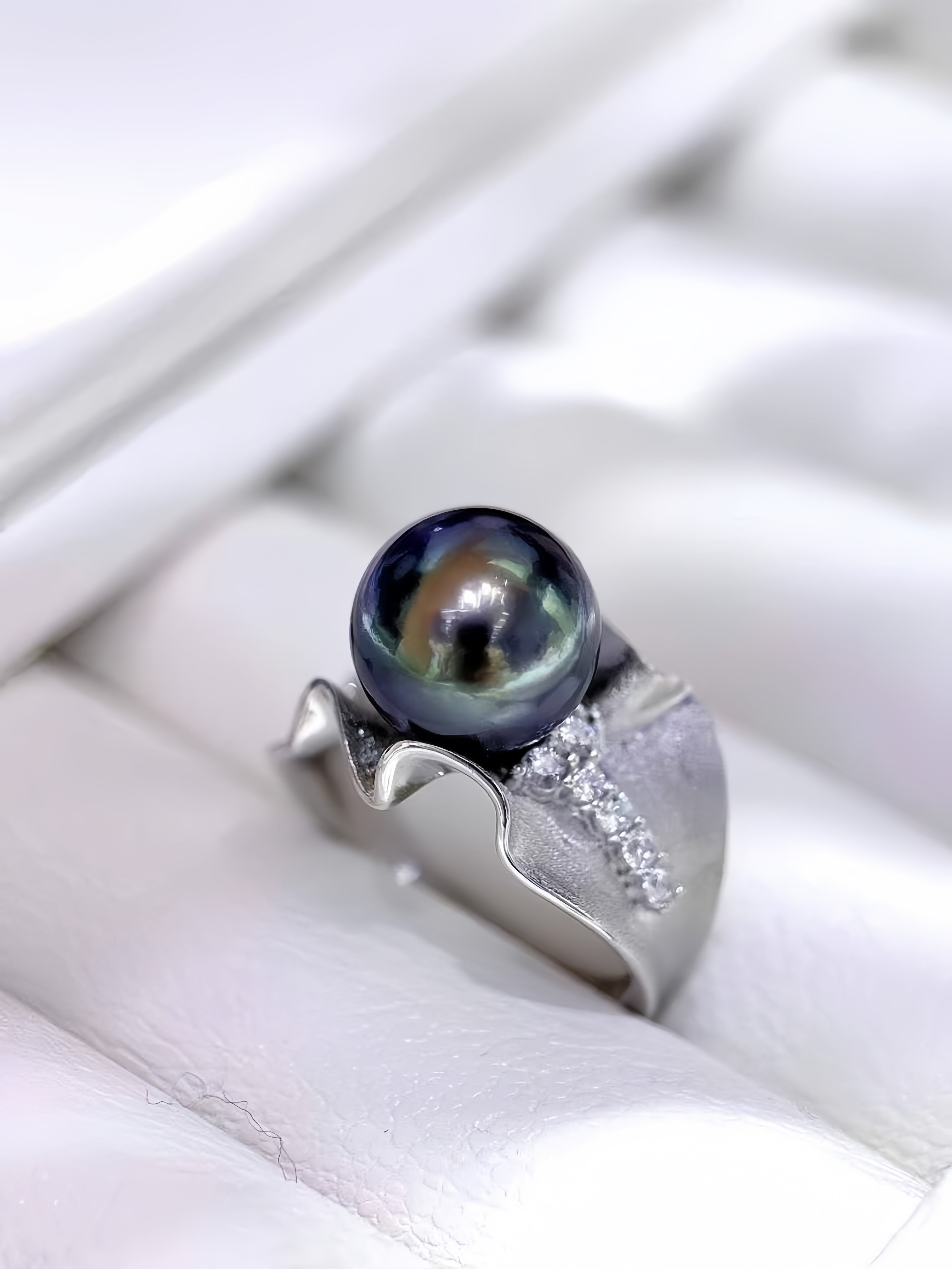 A silver ring featuring a large, round, iridescent dark pearl and a textured, wavy silver band accented with small clear stones, displayed on a white cushion.