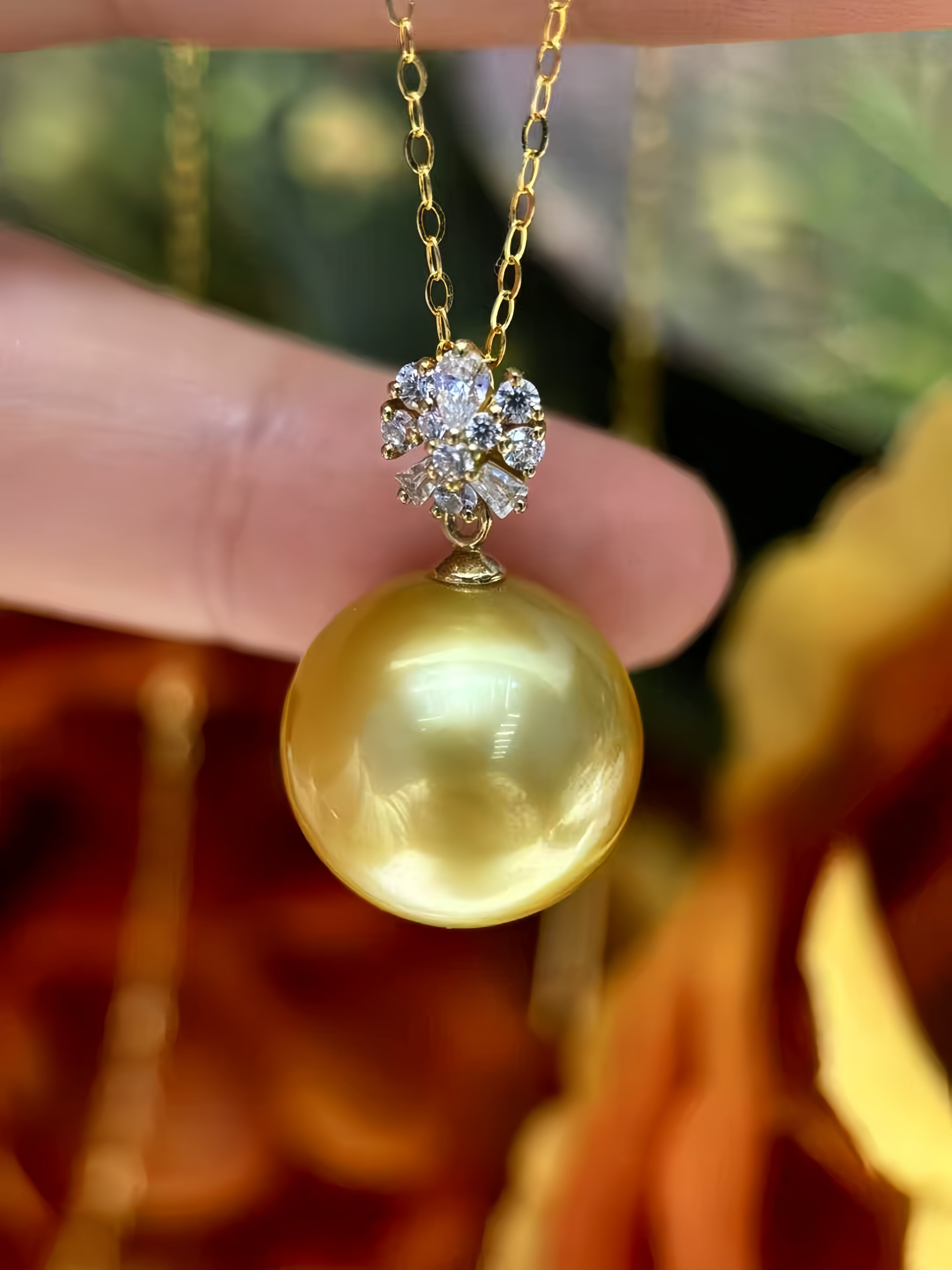 A close-up of a gold pendant necklace featuring a large, lustrous yellow pearl and a diamond-studded floral cluster, held by a hand against a warm, blurred background.