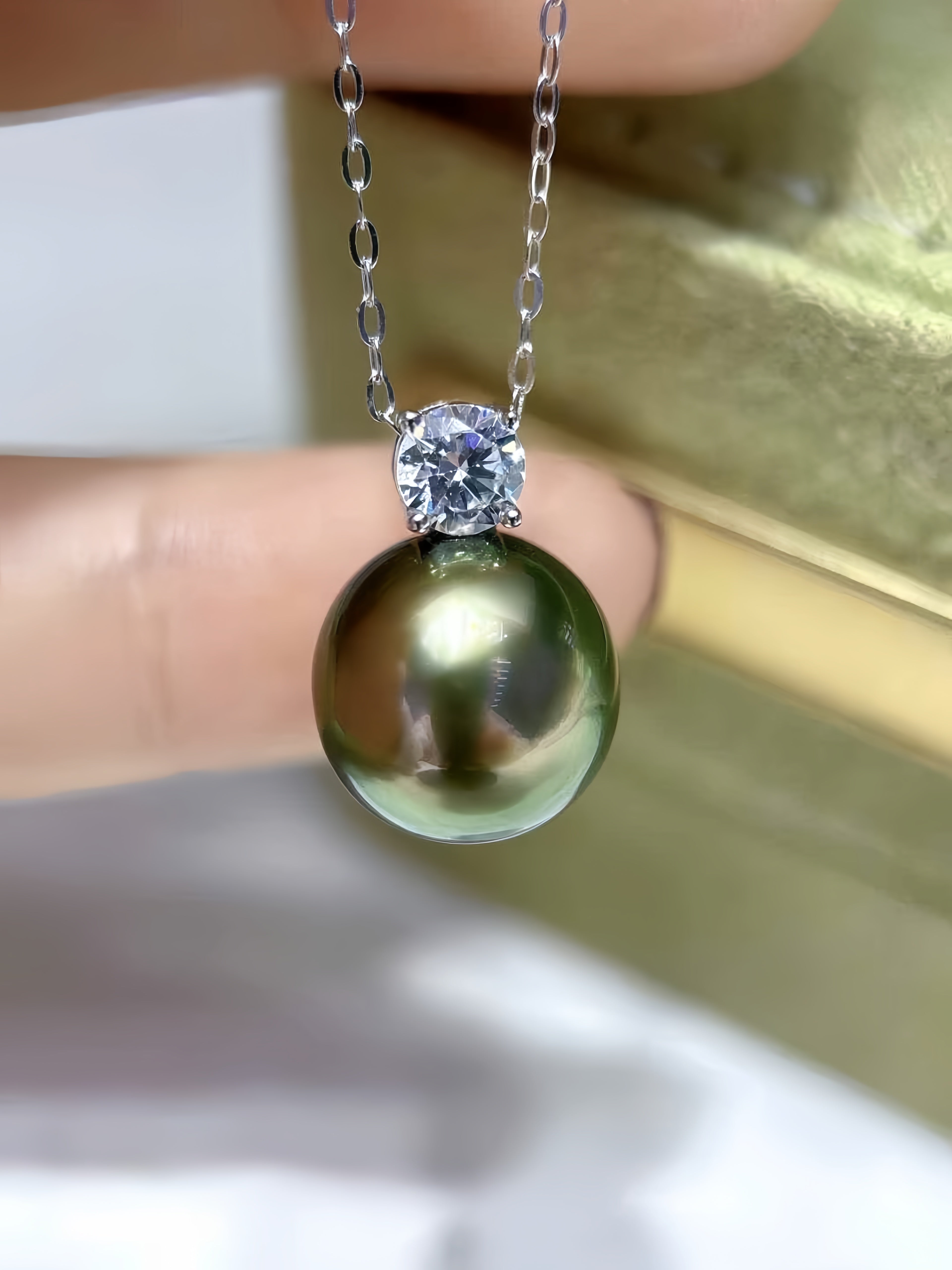 A close-up of a silver chain necklace with a round, dark green-black pearl pendant held by fingers, featuring a clear round gemstone in a prong setting at the top.