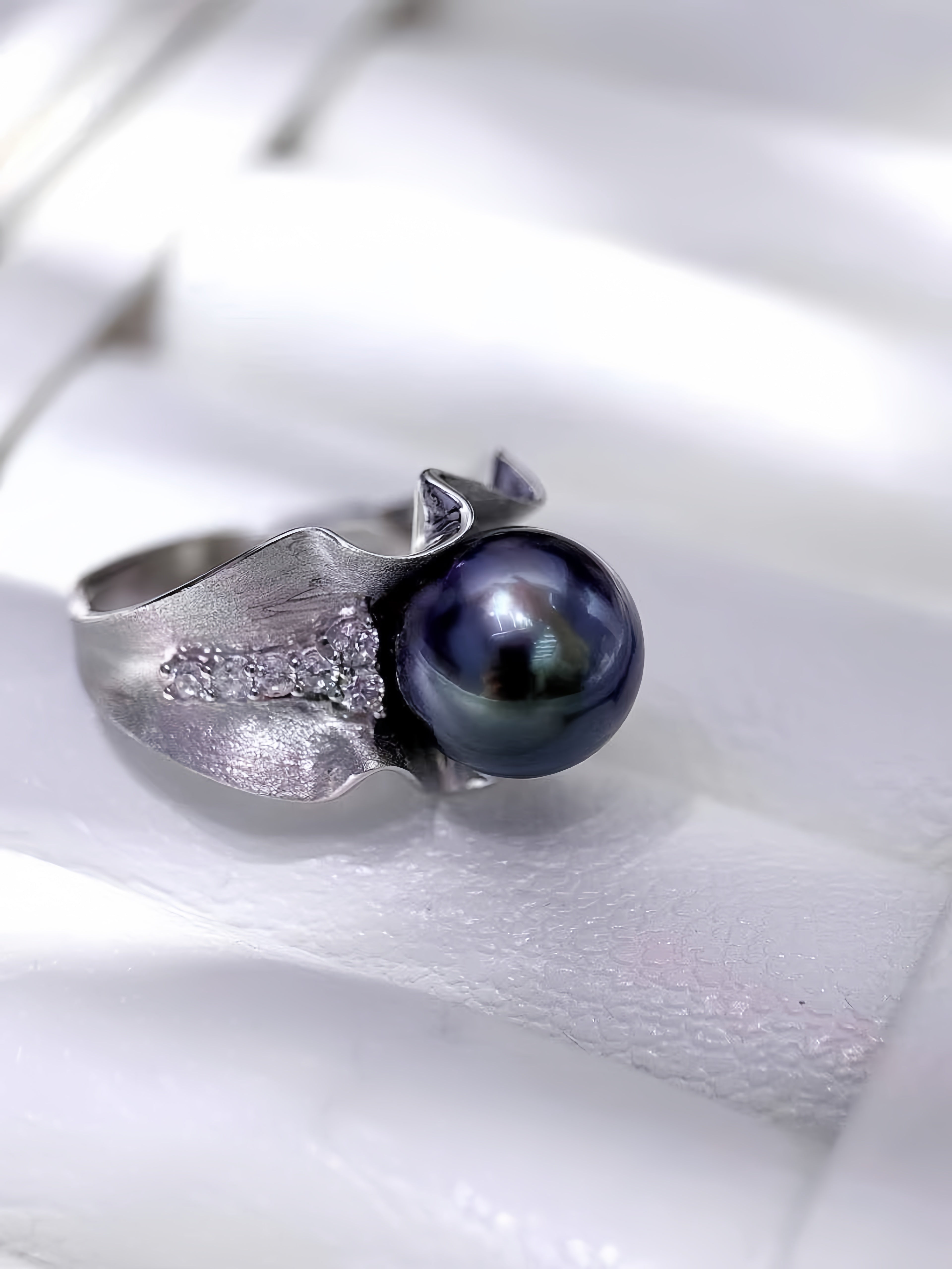A silver ring featuring a large, dark iridescent freshwater pearl and a textured, leaf-like silver setting with small clear stones, displayed on a white textured surface.