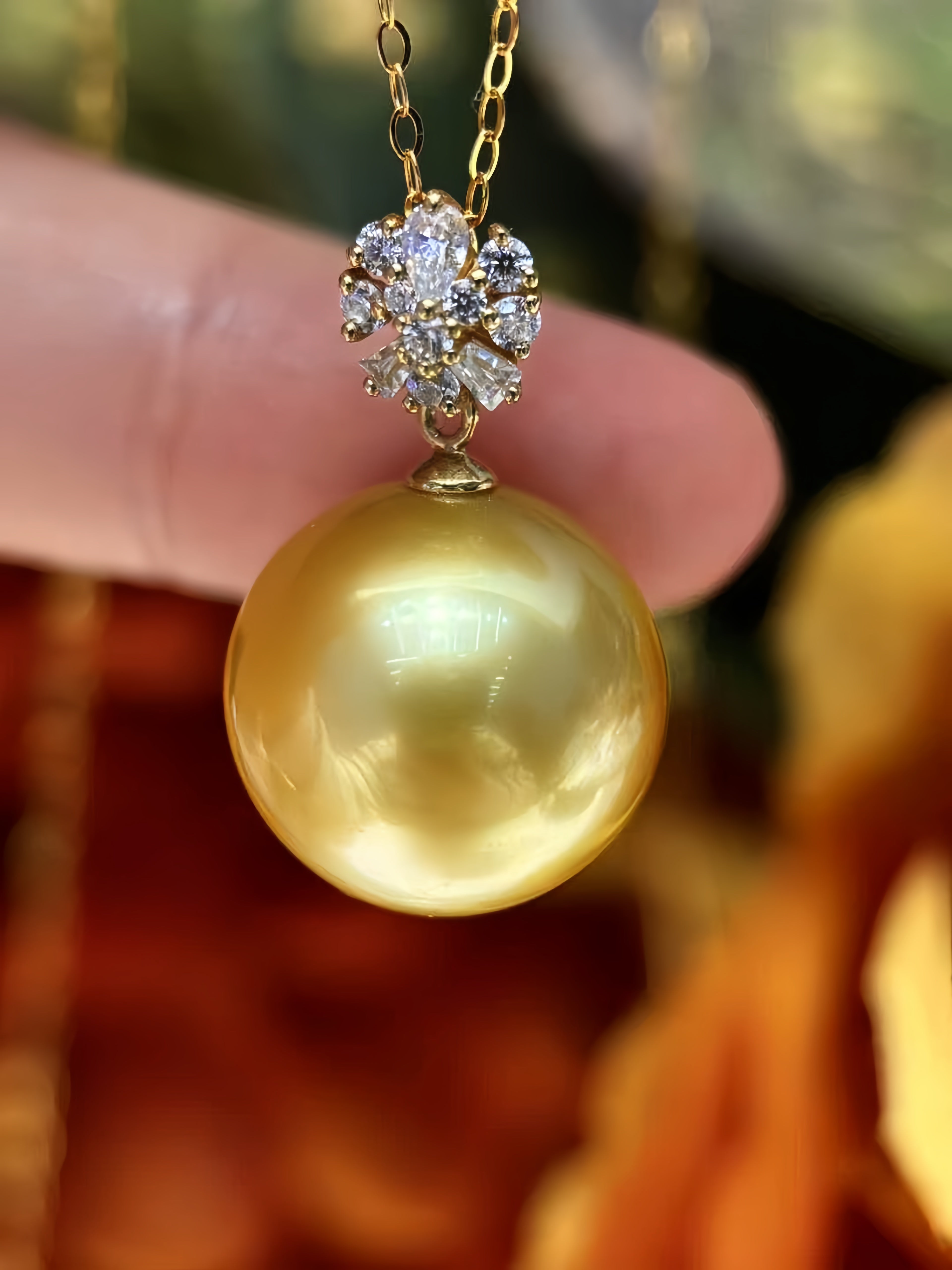 A close-up of a gold pendant necklace featuring a large, lustrous golden pearl and a small cluster of clear stones, held by a person's fingers against a warm, blurred background.