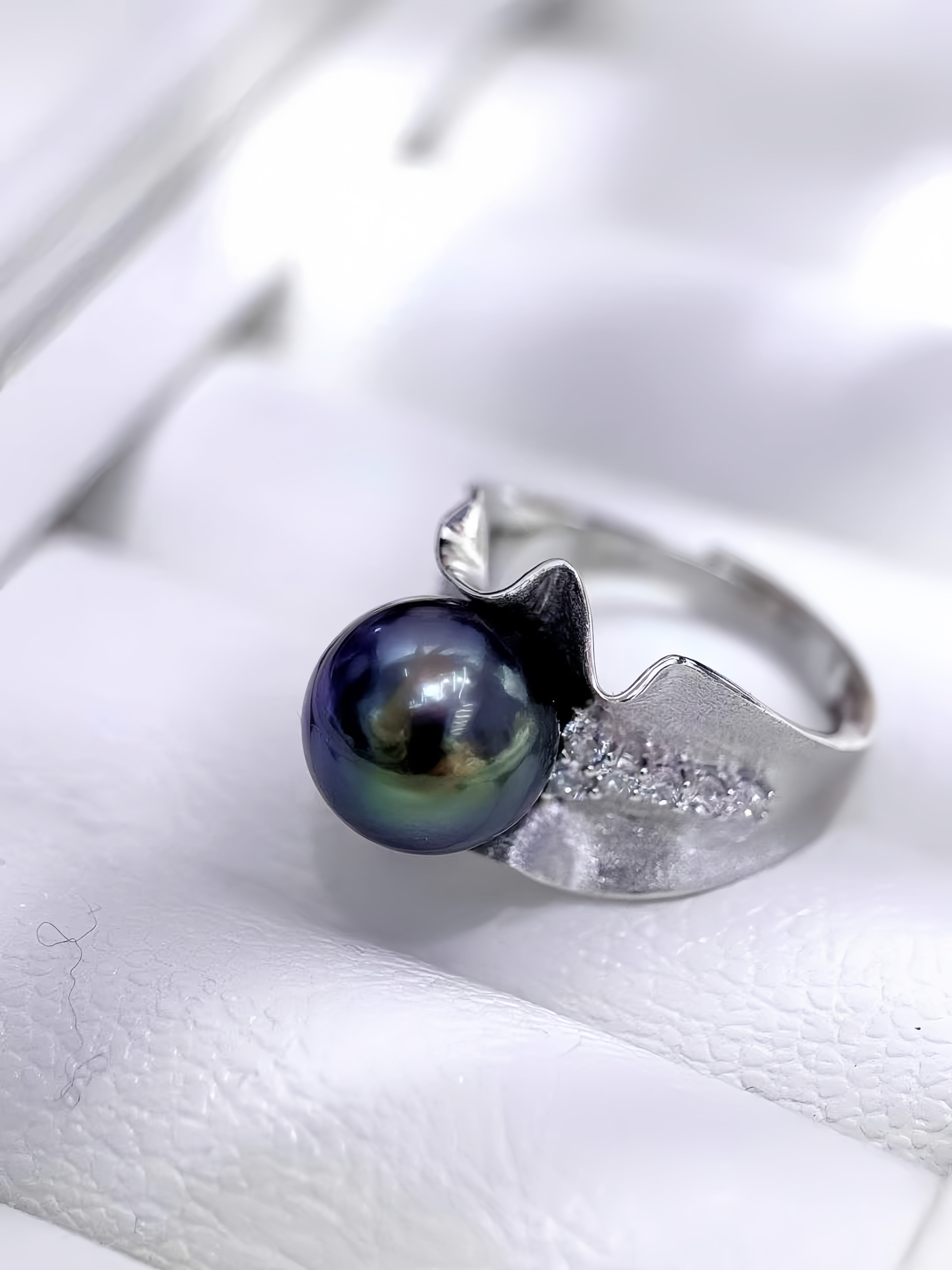 A silver ring featuring a large, dark iridescent pearl and a textured silver leaf-shaped setting with small clear stones, displayed on a white textured surface.