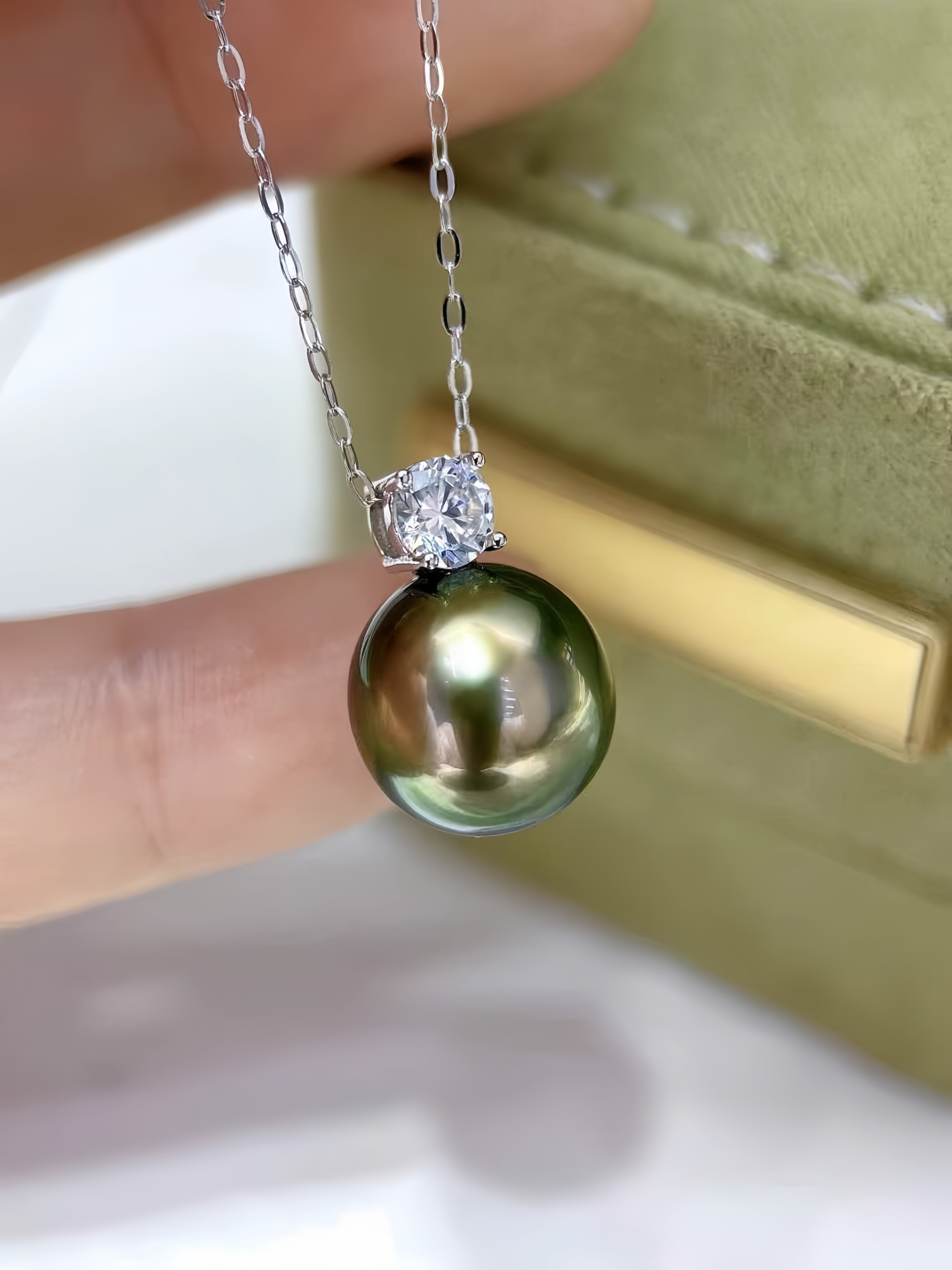 A close-up of a silver pendant necklace featuring a round green-black Tahitian pearl and a clear round-cut gemstone, held by a hand in front of a green jewelry box.