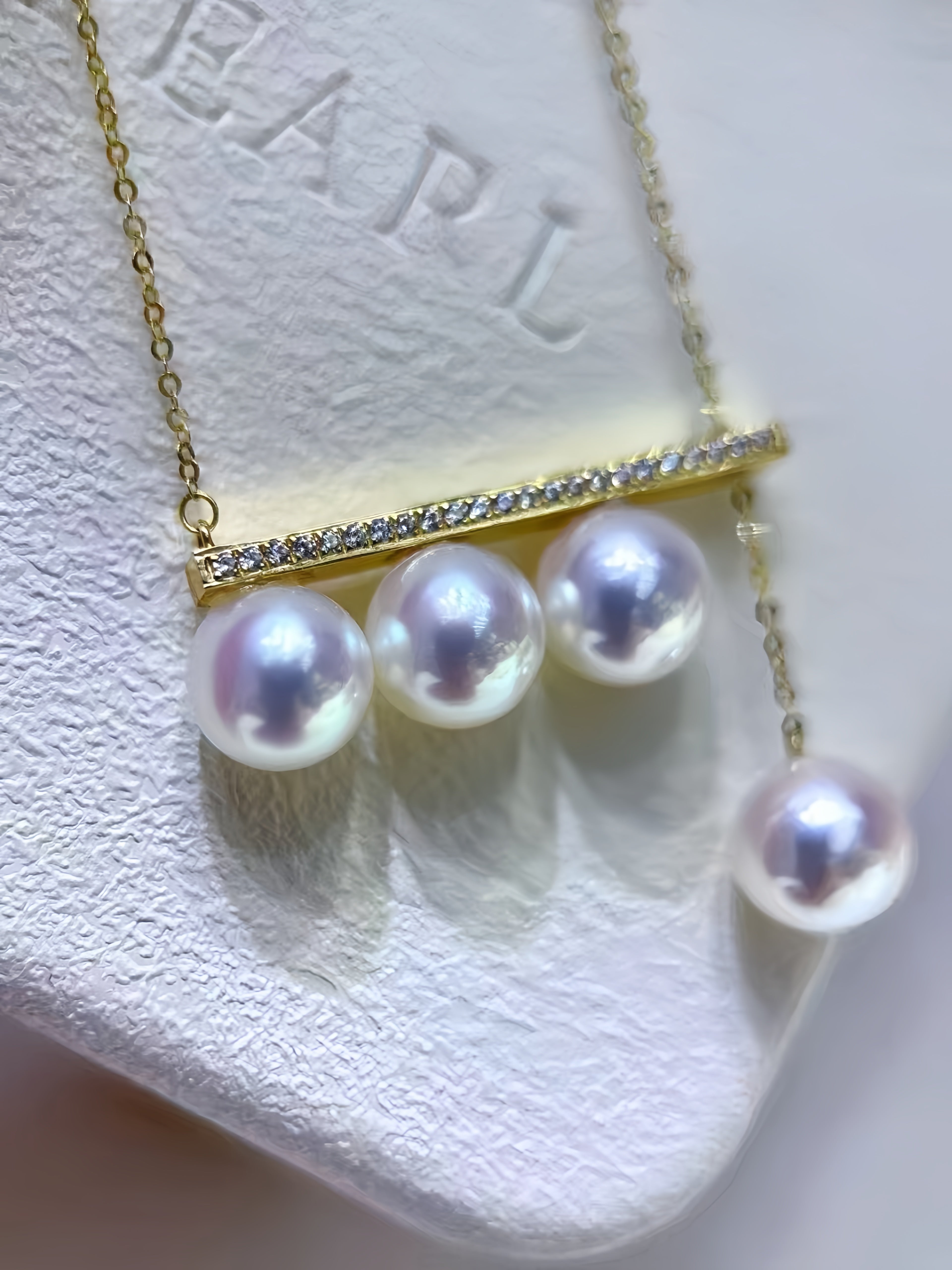 A gold bar necklace with three large white freshwater pearls and a dangling pearl drop, displayed on a textured white jewelry stand.