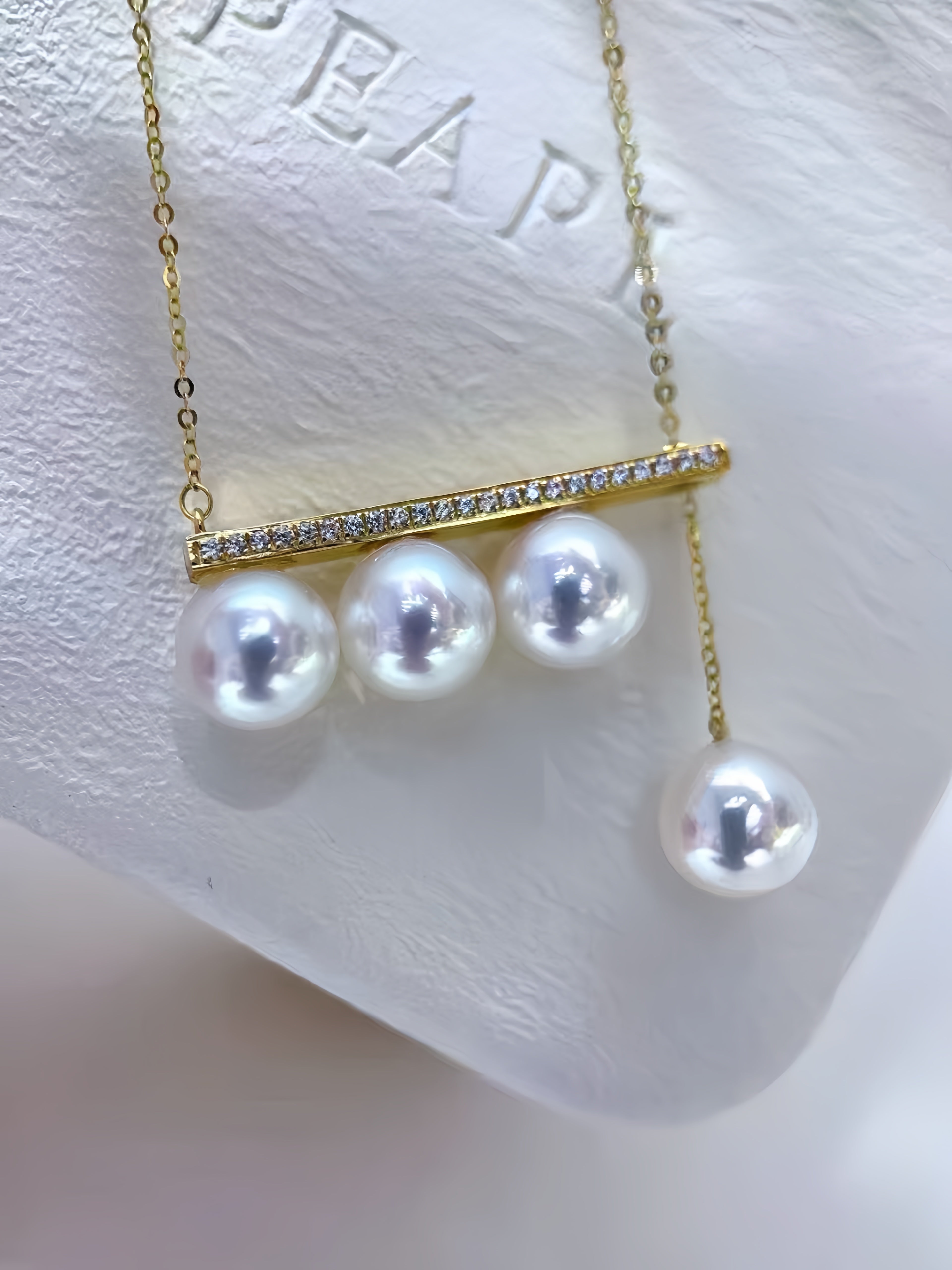 A gold necklace with a horizontal bar set with small clear stones and three dangling white pearls, displayed on a white textured jewelry stand.