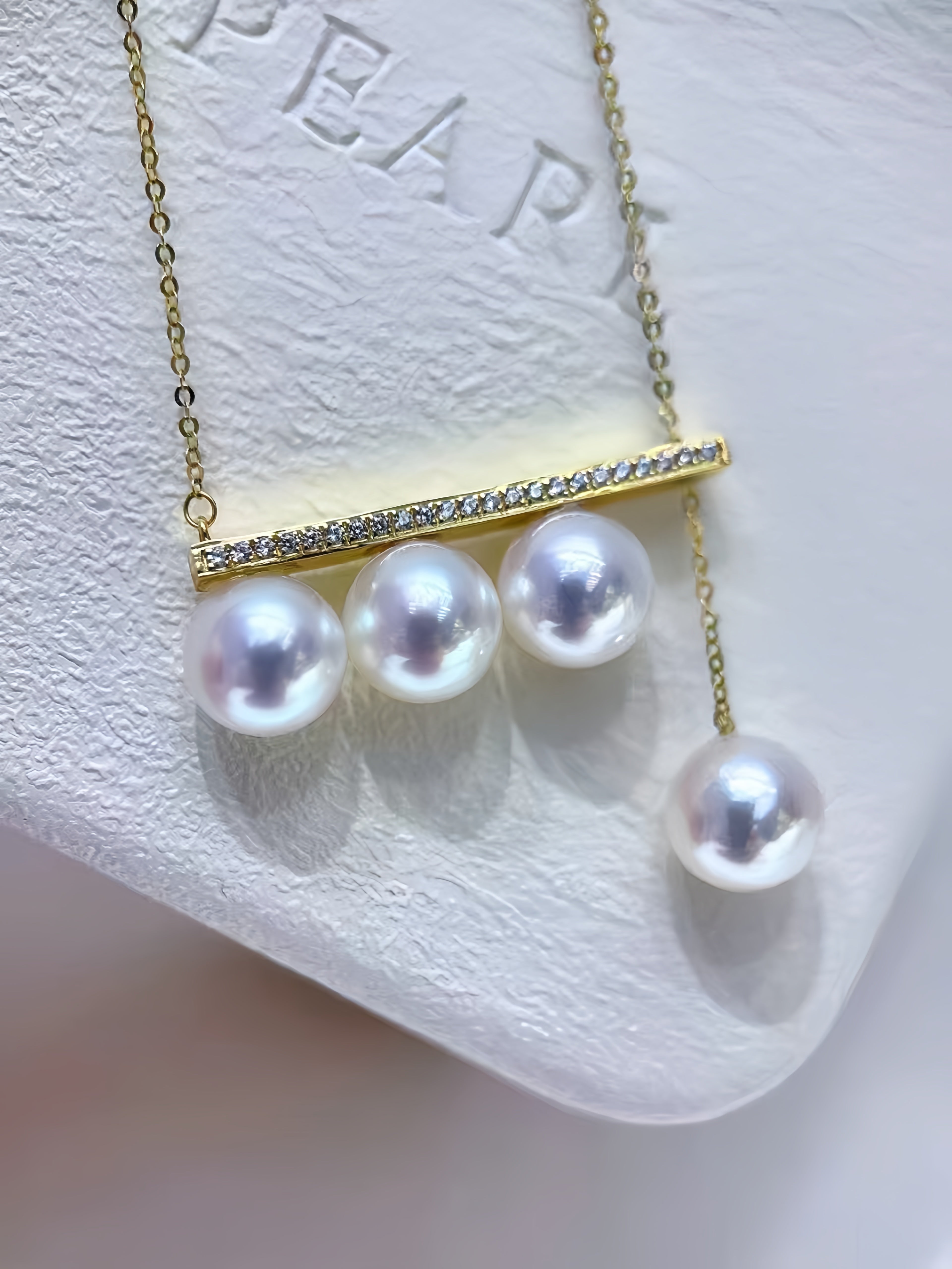 A gold necklace with a horizontal bar set with small clear stones and three dangling white pearls, displayed on a textured white jewelry stand.
