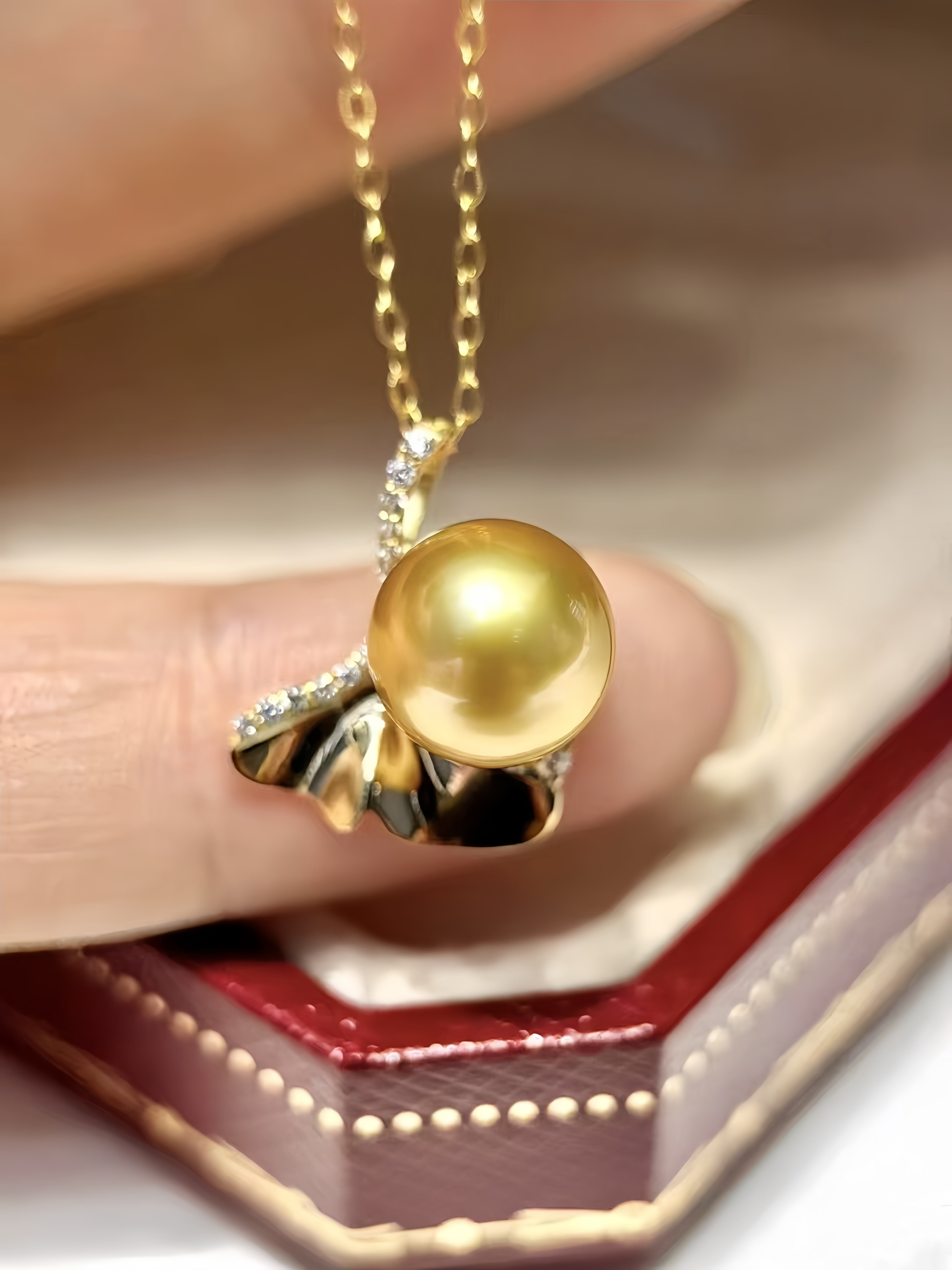 A gold pendant necklace with a large round yellow pearl and a sculpted gold leaf design, held between fingers above an open red jewelry box.