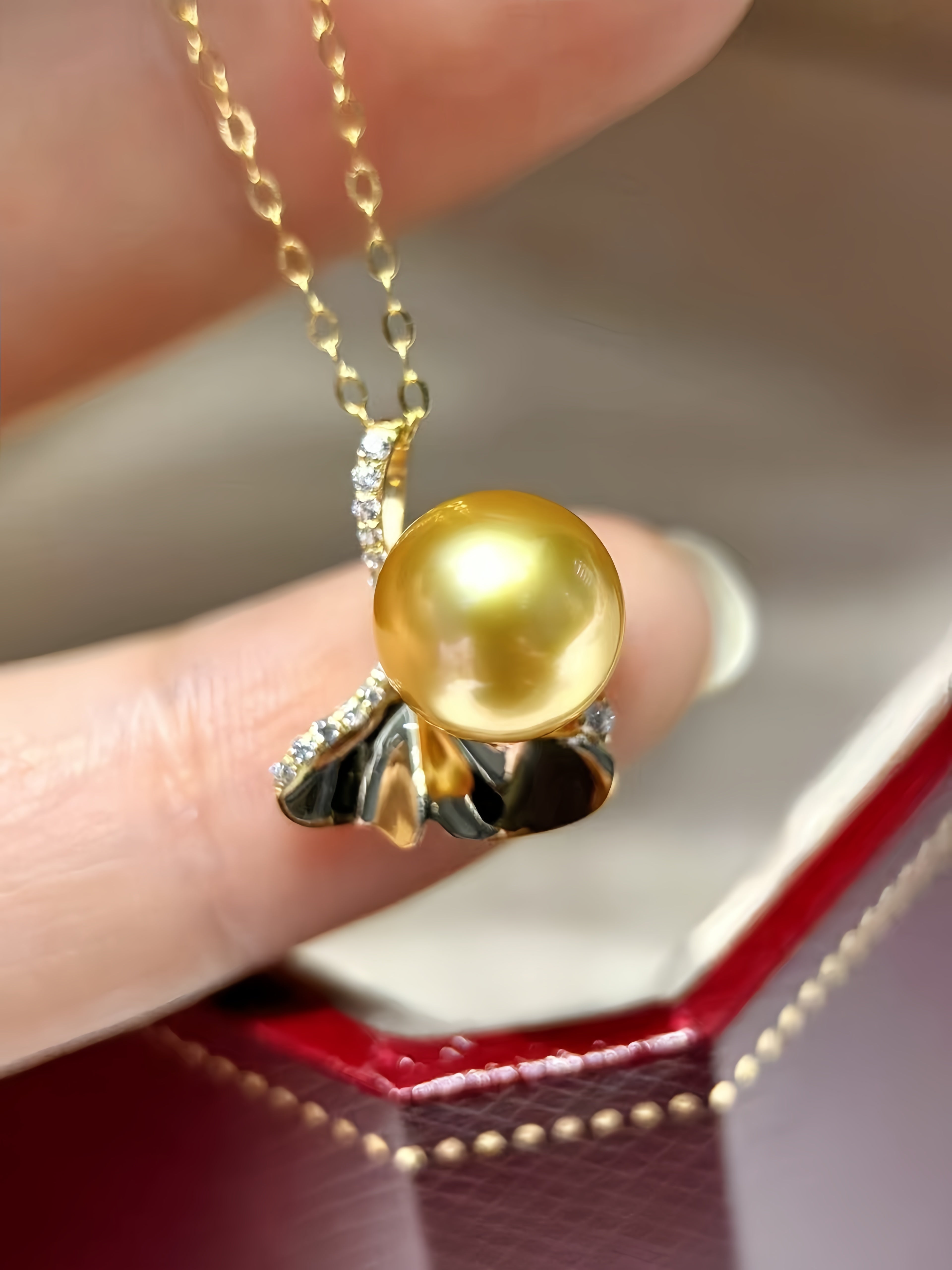 A close-up of a gold pendant necklace with a large round yellow pearl and small clear stones, held between fingers above an open red jewelry box.
