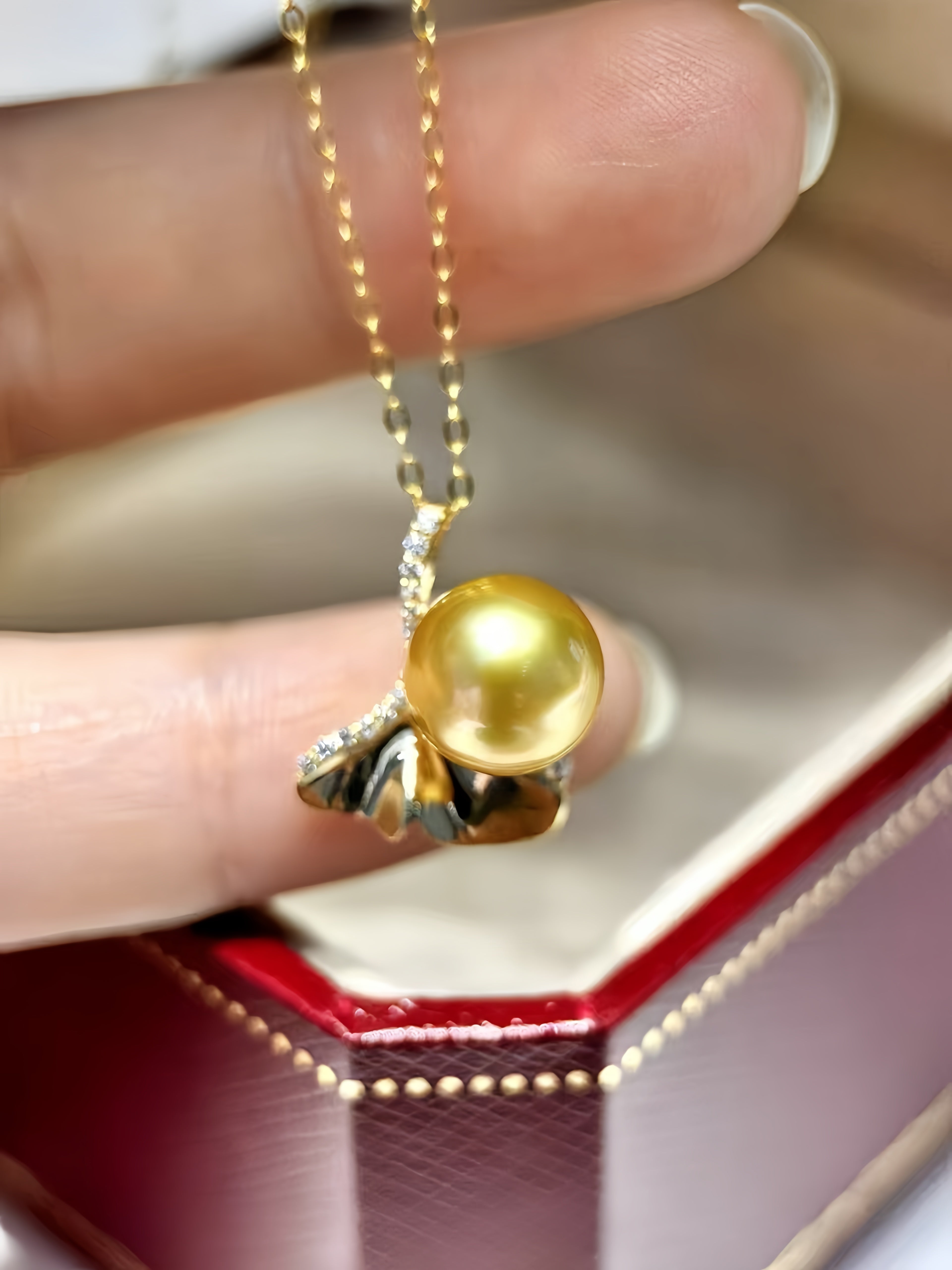 A gold pendant necklace with a round yellow gold pearl and textured gold leaf details, held between fingers above an open red jewelry box.
