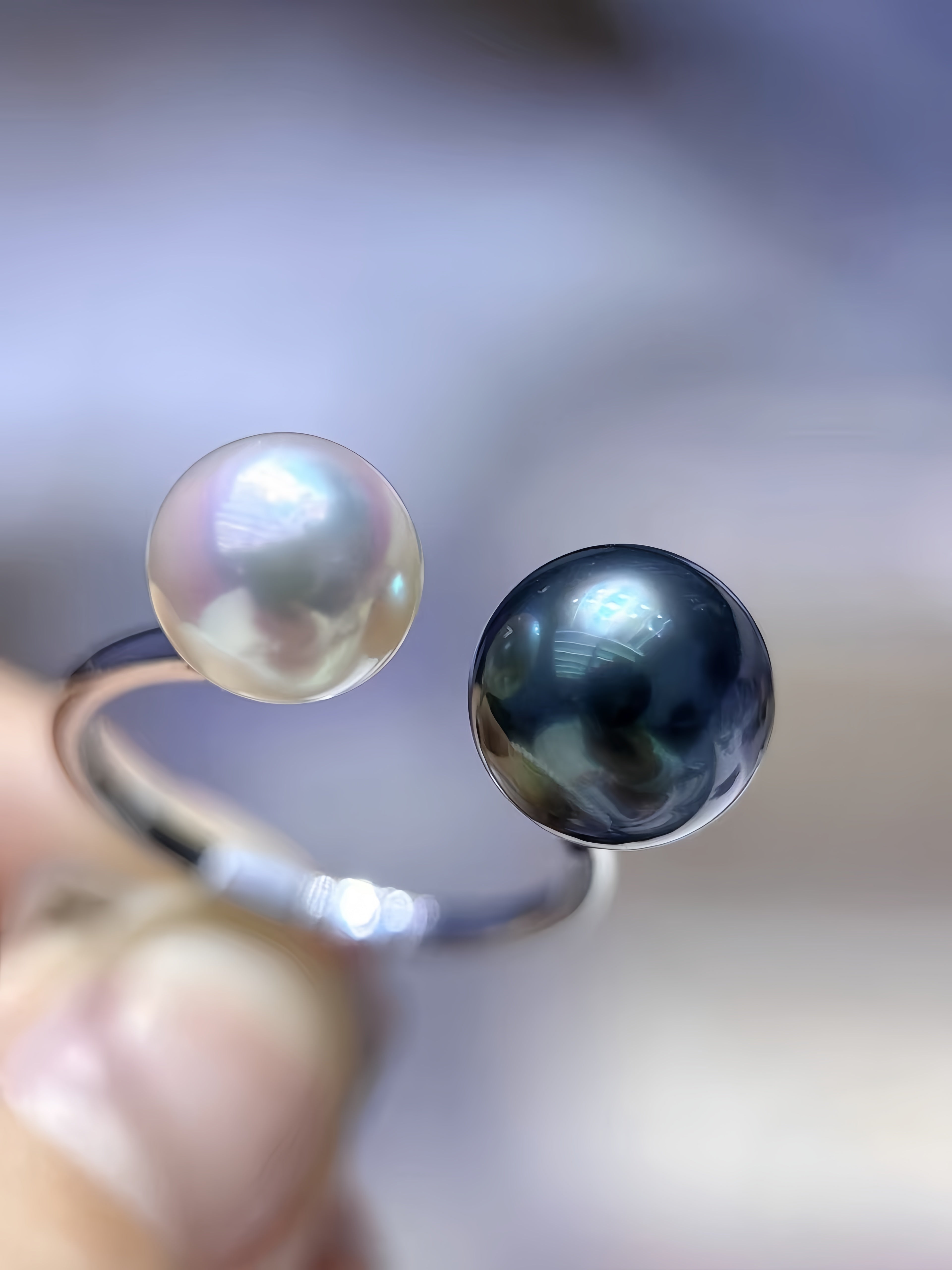 A close-up of a silver-toned ring held by fingers, featuring two round pearls: one lustrous white pearl and one dark black pearl.