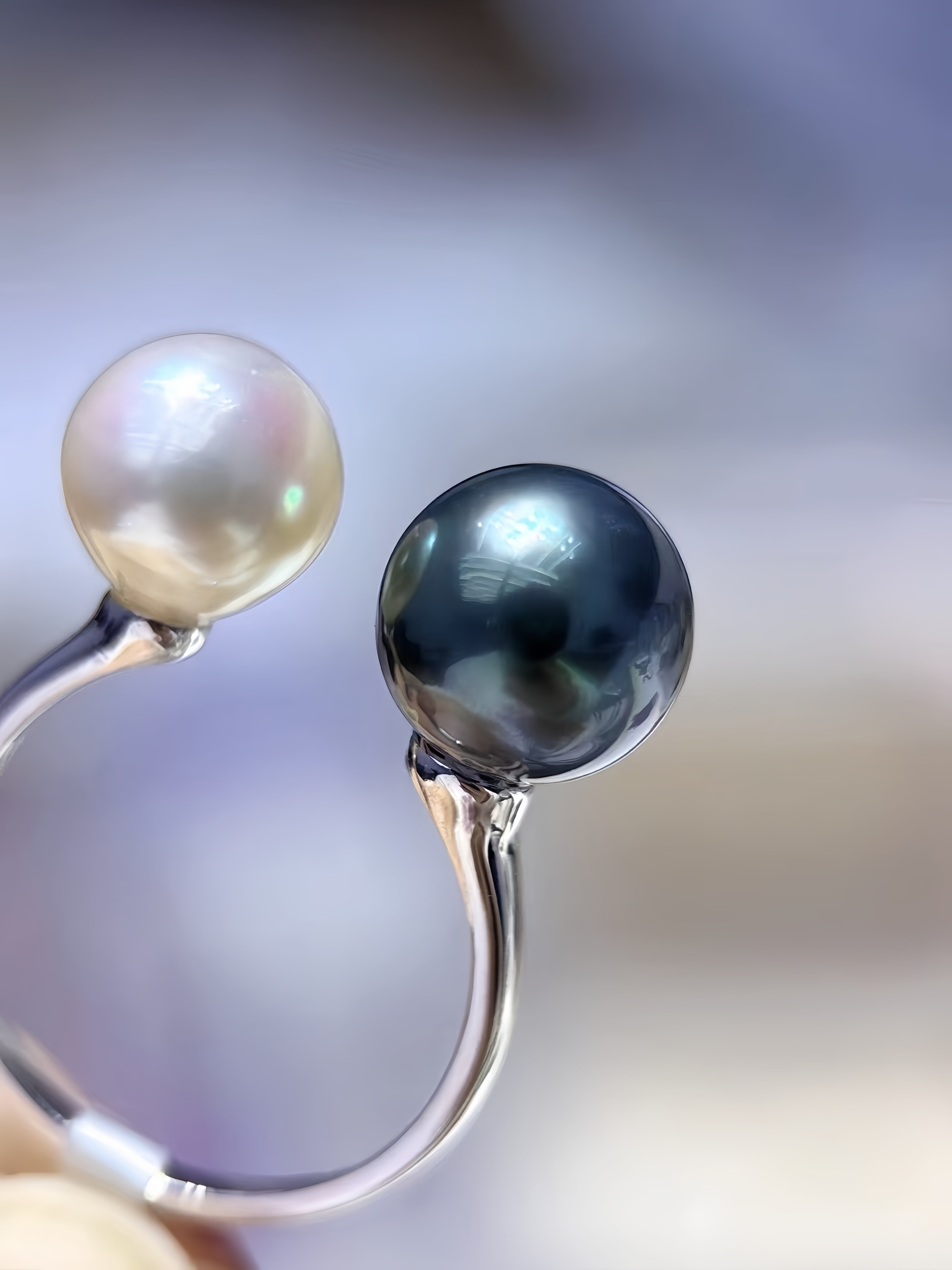 A close-up of a silver ring with two round pearls, one lustrous white pearl and one dark gray-black pearl, set against a soft blurred background.