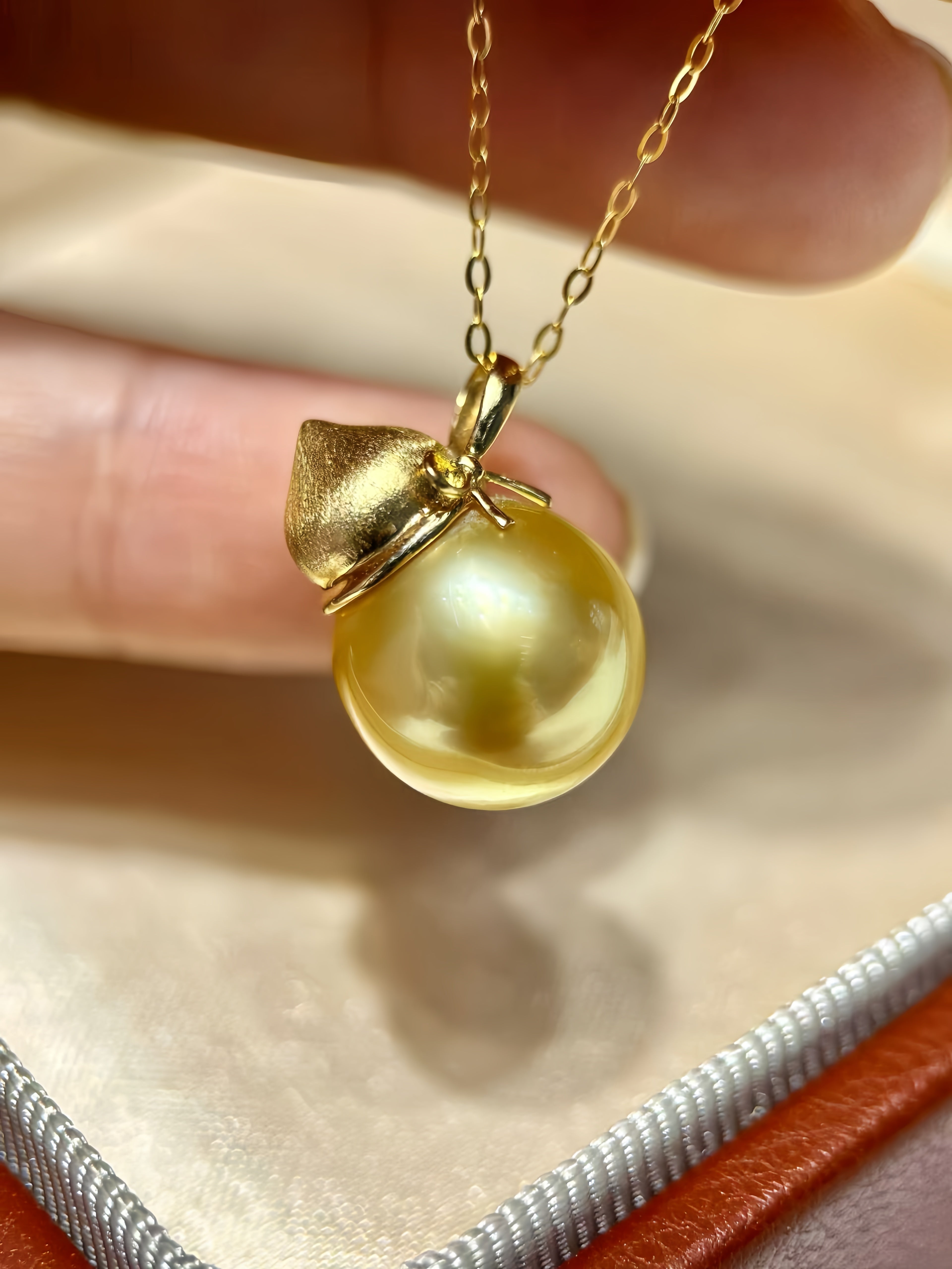 A gold pendant necklace featuring a large, lustrous yellow pearl pendant held by a hand, set against a soft beige satin-lined box.