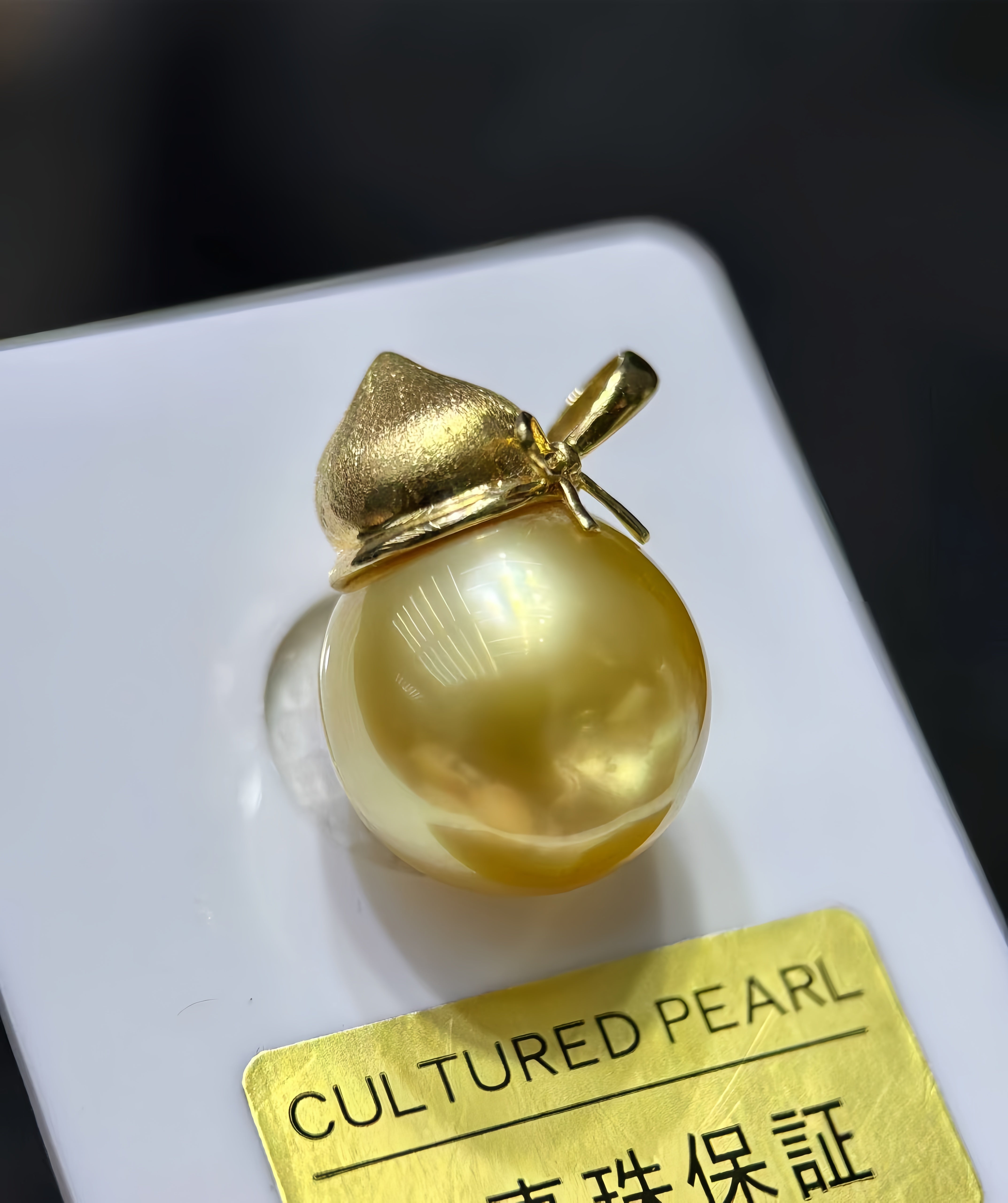 A close-up of a cultured pearl pendant with a gold cap, displayed on a white card with a gold label that reads 'CULTURED PEARL'.