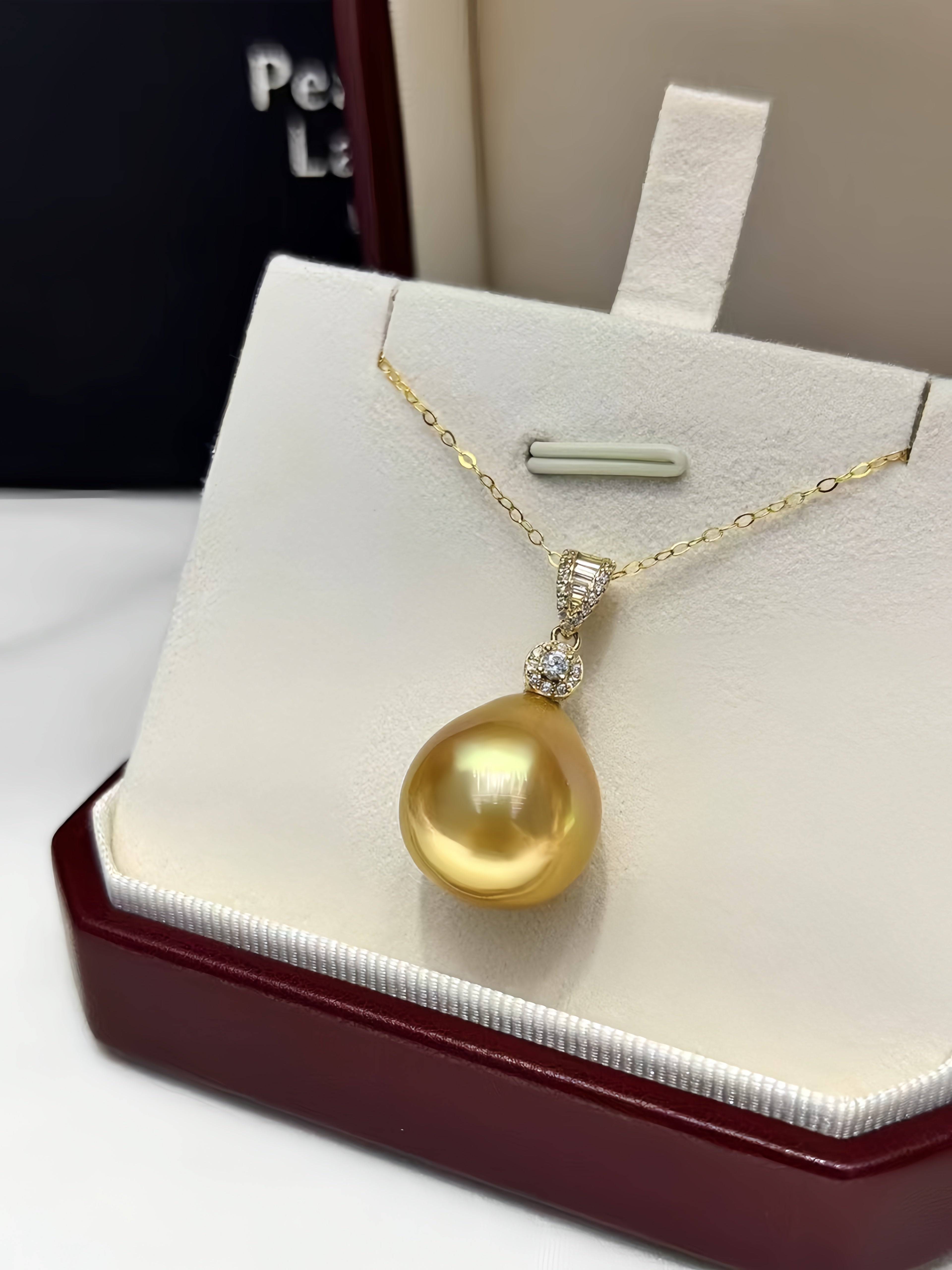 A gold pendant necklace featuring a large, lustrous baroque-style gold pearl, displayed on a white jewelry stand inside a dark red presentation box.