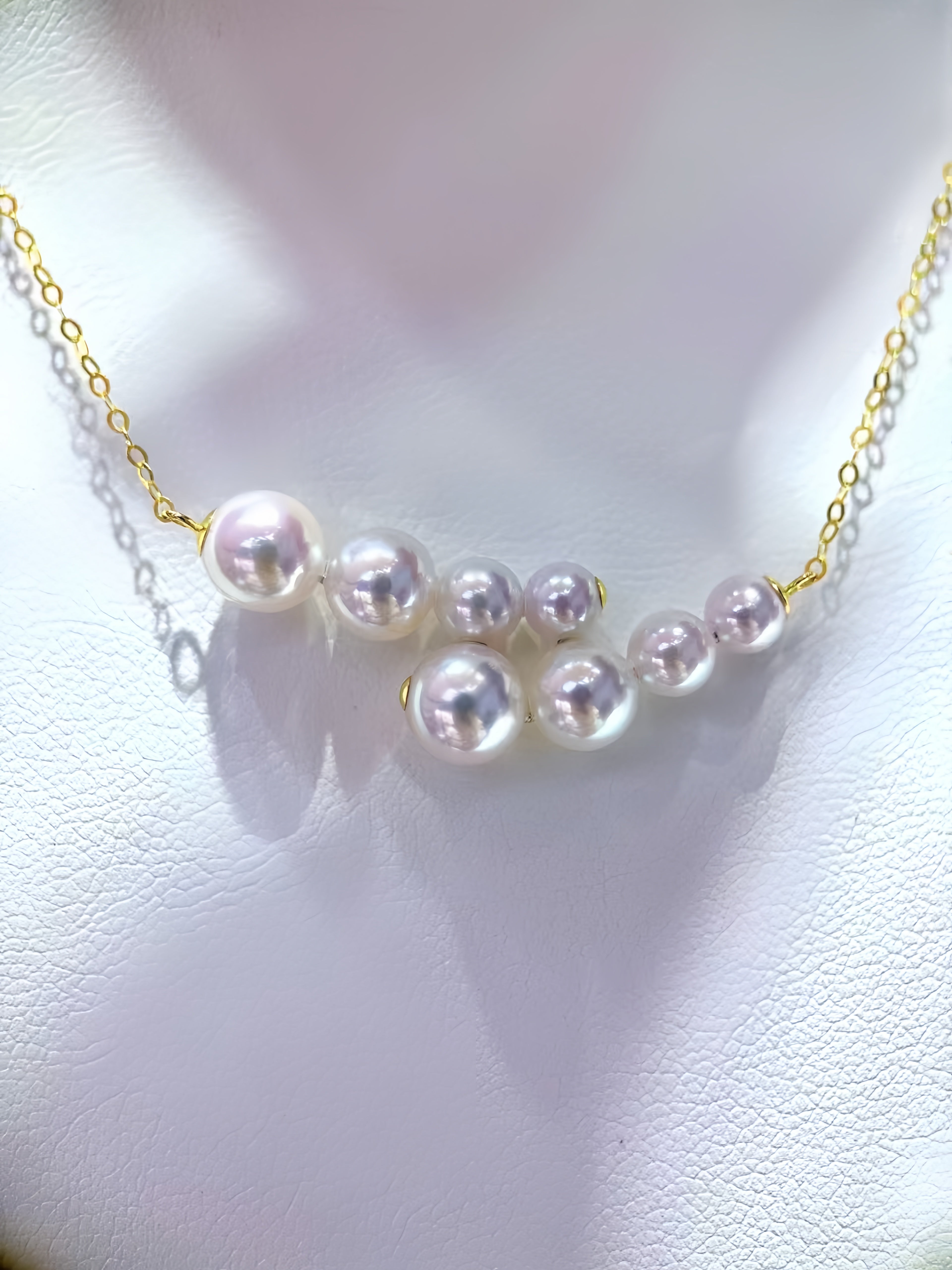 A gold chain necklace featuring a curved arrangement of white freshwater pearls, displayed on a textured white surface.